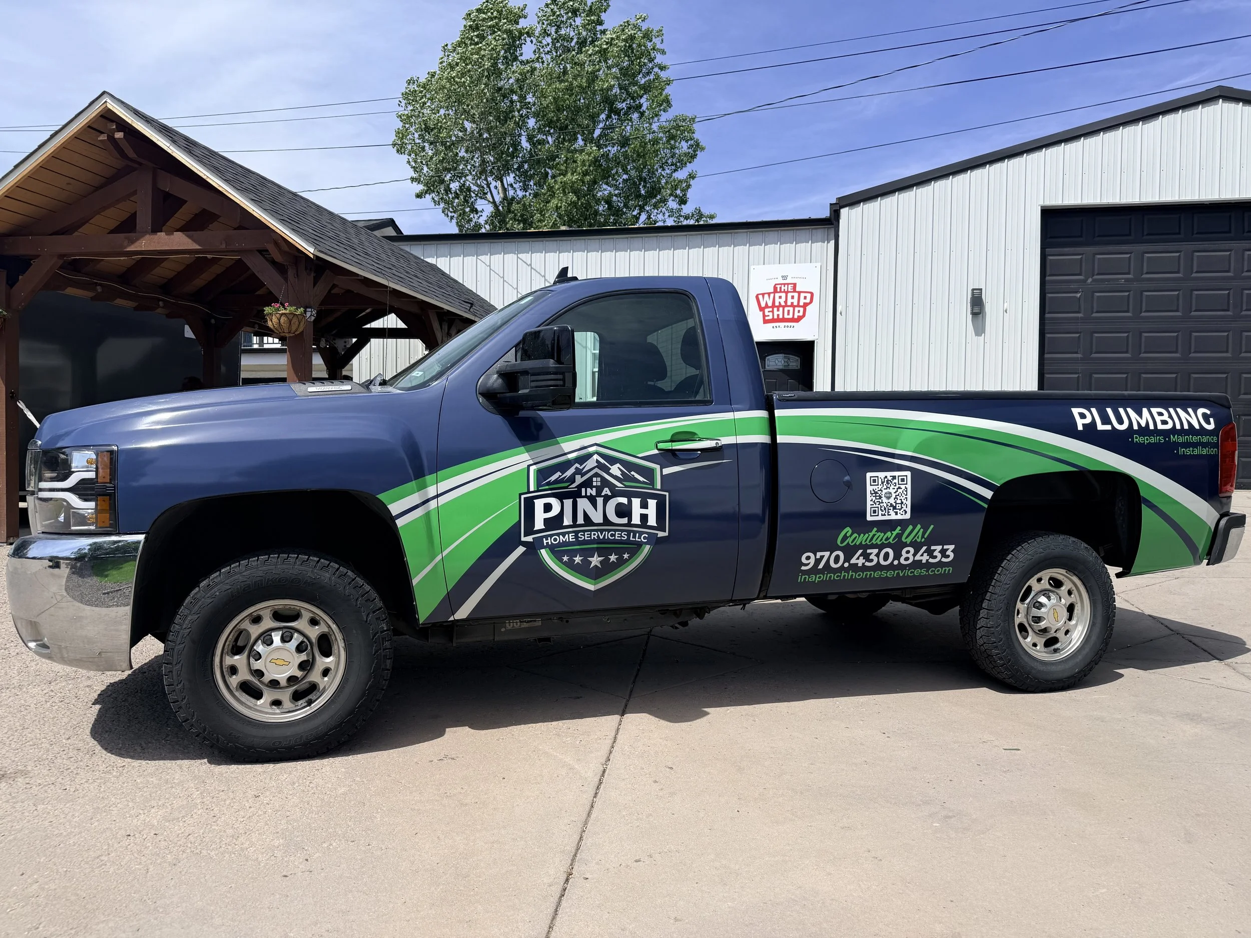 A blue pickup truck with green and black graphics parked outdoors in front of a metal building with a sign that reads "The Wrap Shop". Behind, there is a large tree and power lines.
