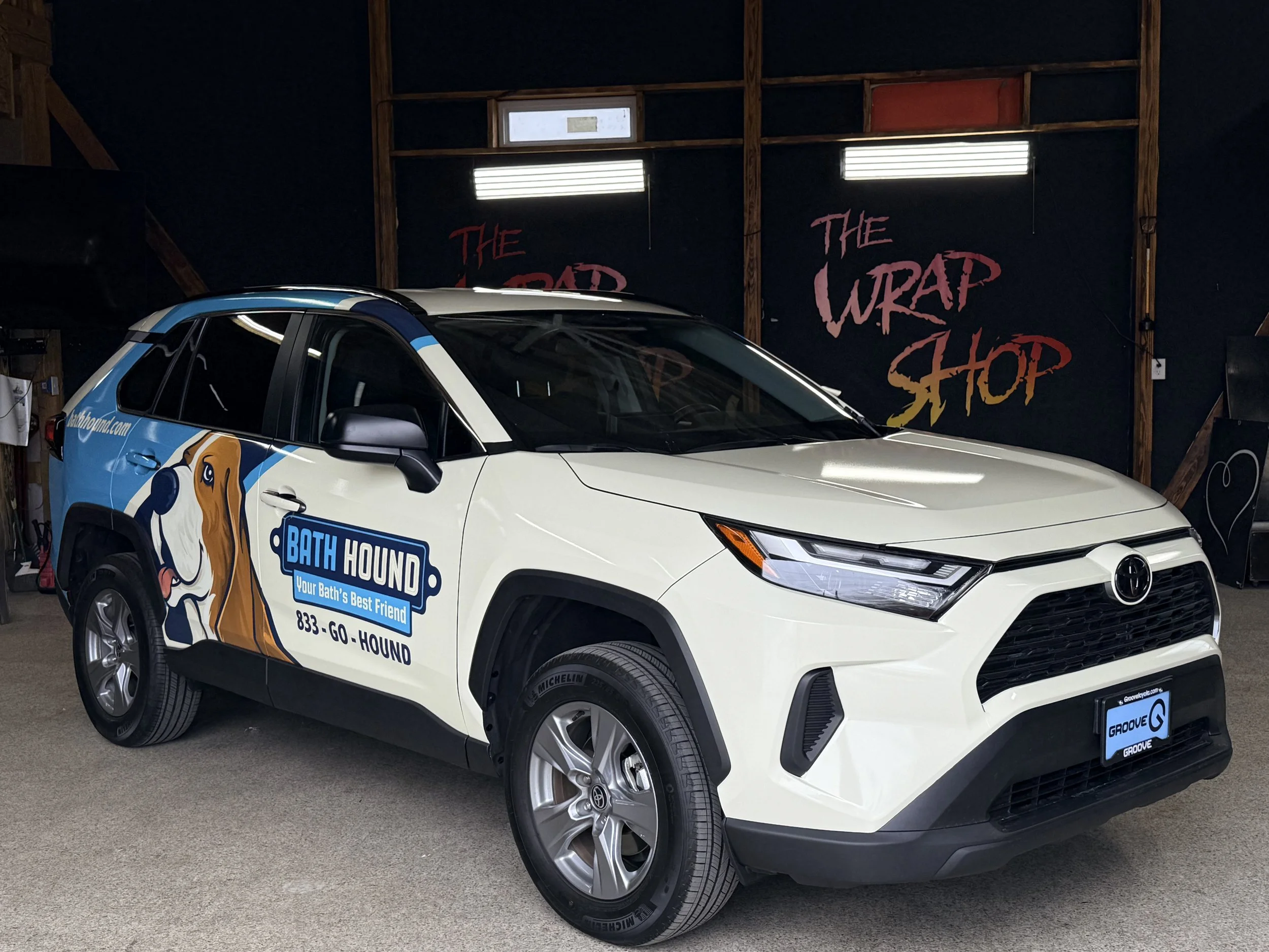 White and black Toyota vehicle with branding for Bath Hound, a pet grooming service, parked inside a garage with dark walls and wooden framing. The garage wall has a black background with the words 'The Wrap Shop' painted in red and pink letters.