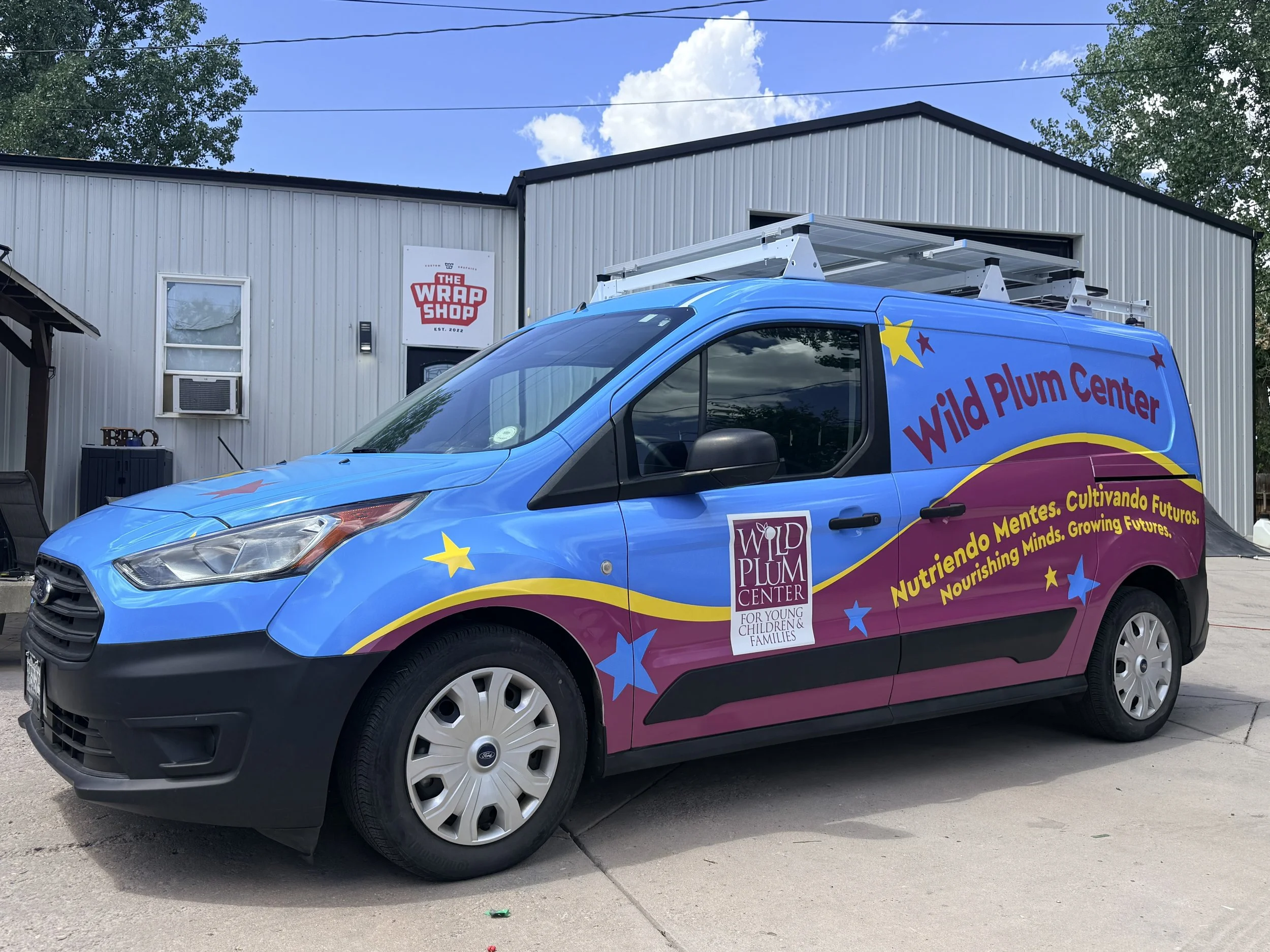 A blue and purple van with colorful star graphics and signage for Wild Plum Center. The van is parked outside a building with a sign that reads 'The Wrap Shop'. The sky is mostly clear with some clouds, and there are trees in the background.