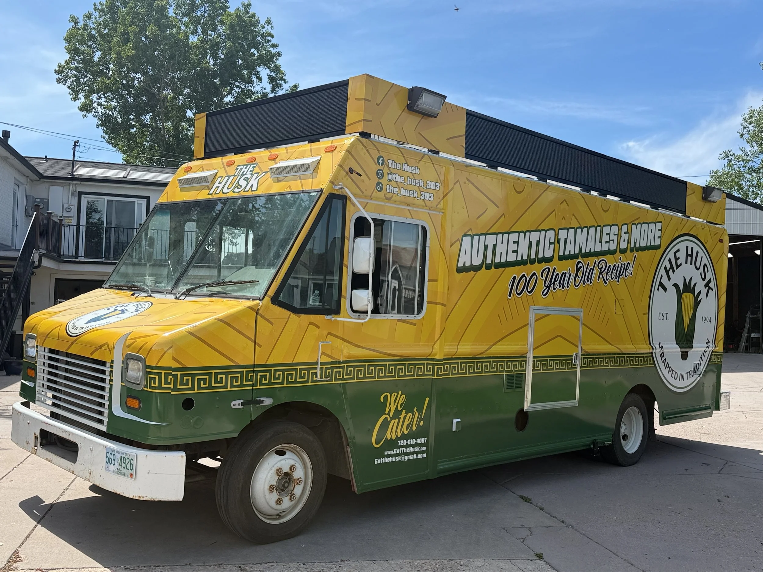Yellow and green food truck named 'The Husk' parked on a paved area in front of residential buildings with trees in the background. The truck advertises authentic tamales and mentions a 100-year-old recipe.