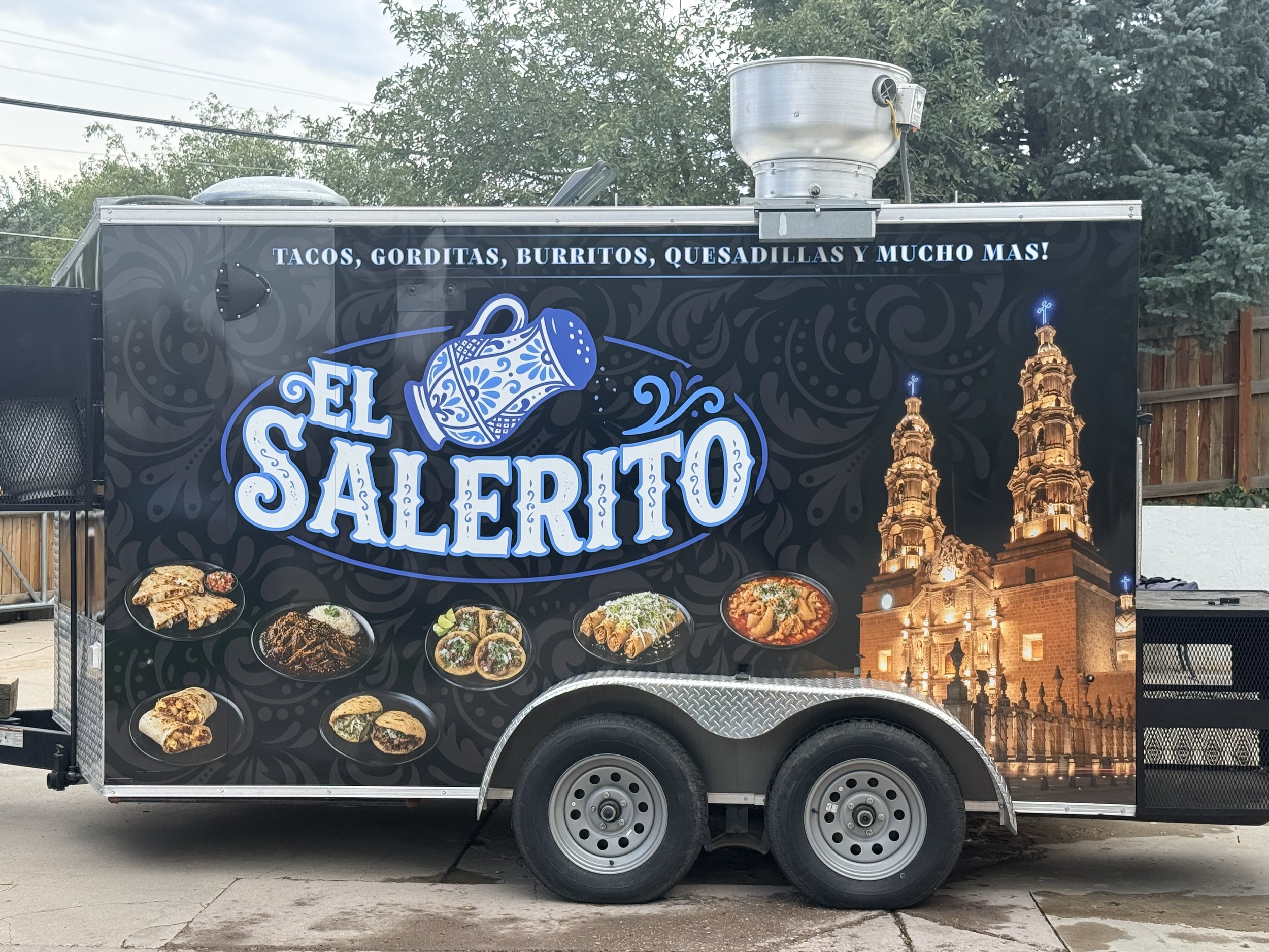 A food truck with a sign advertising tacos, gorditas, burritos, quesadillas, and more in Spanish. The truck has images of various Mexican dishes and features a background that includes a lit structure resembling the Cathedral of Our Lady of Guadalupe