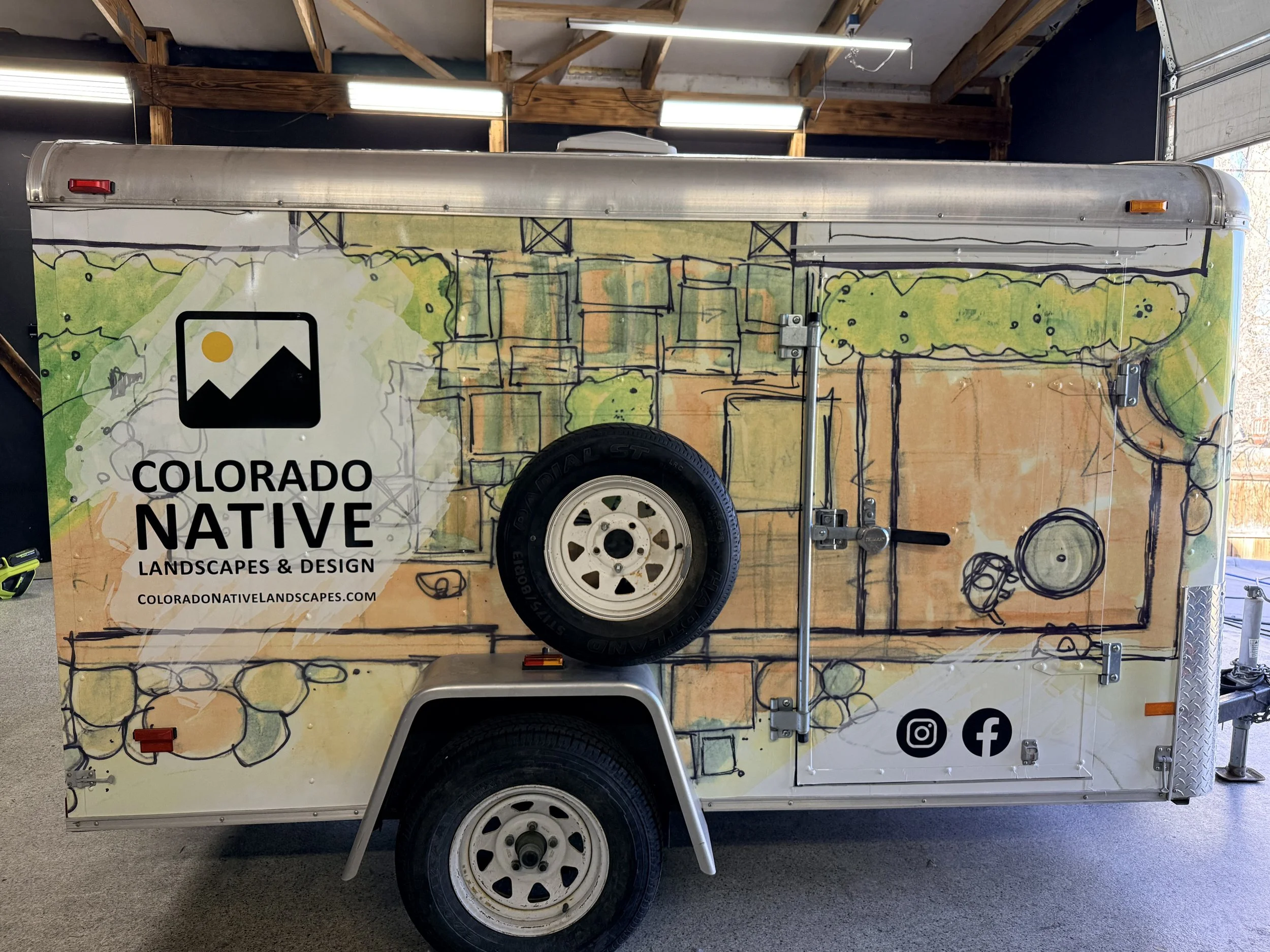 A trailer with a colorful landscape illustration and the words 'Colorado Native Landscapes & Design.' The trailer features a hand-drawn style illustration of trees, landscapes, and infrastructure like bridges. It has a tire and wheel at the bottom, w