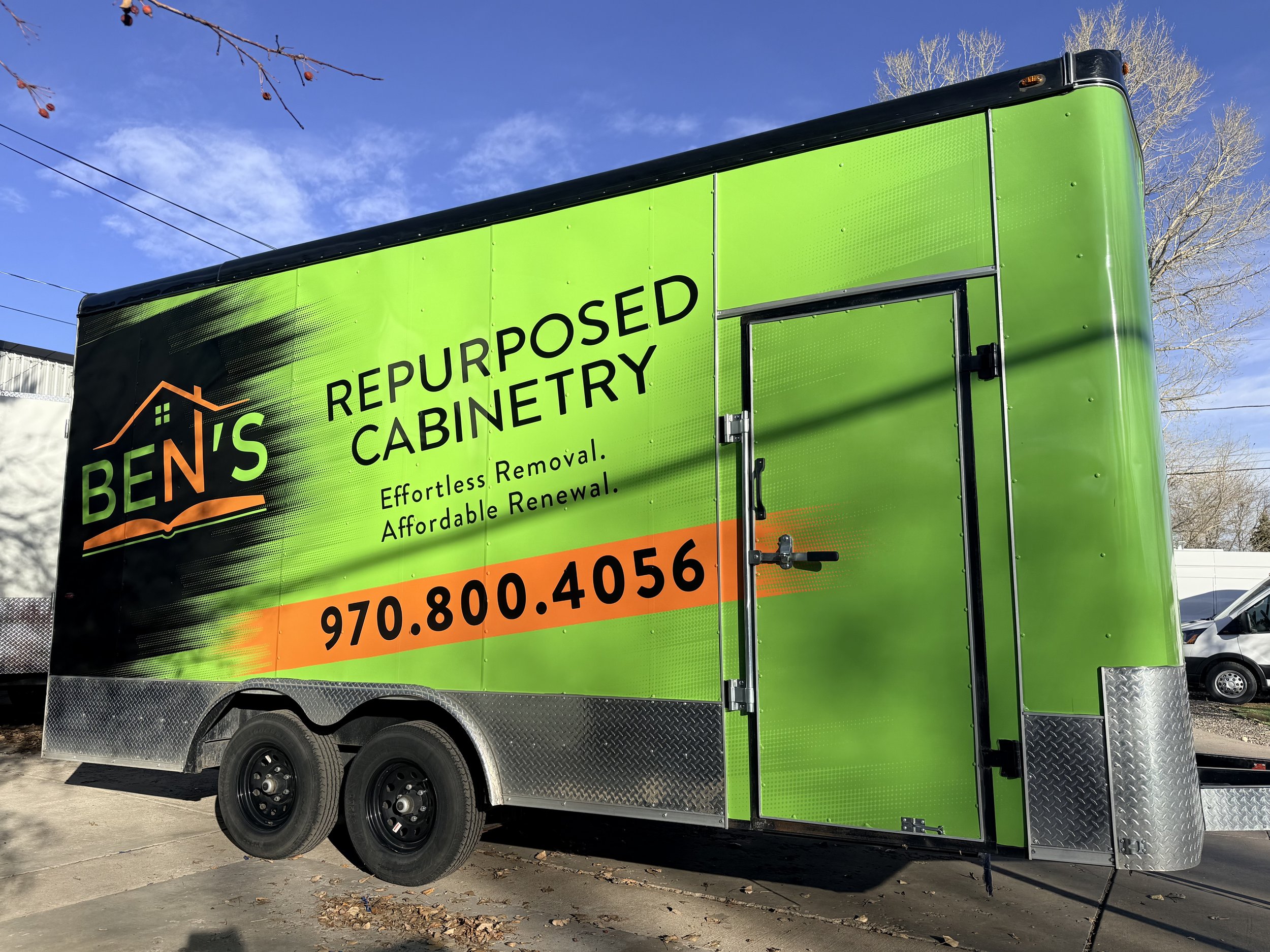 Green mobile unit with black and orange signage advertising residential re-purposing services, parked on a street with a clear blue sky and bare trees in the background.