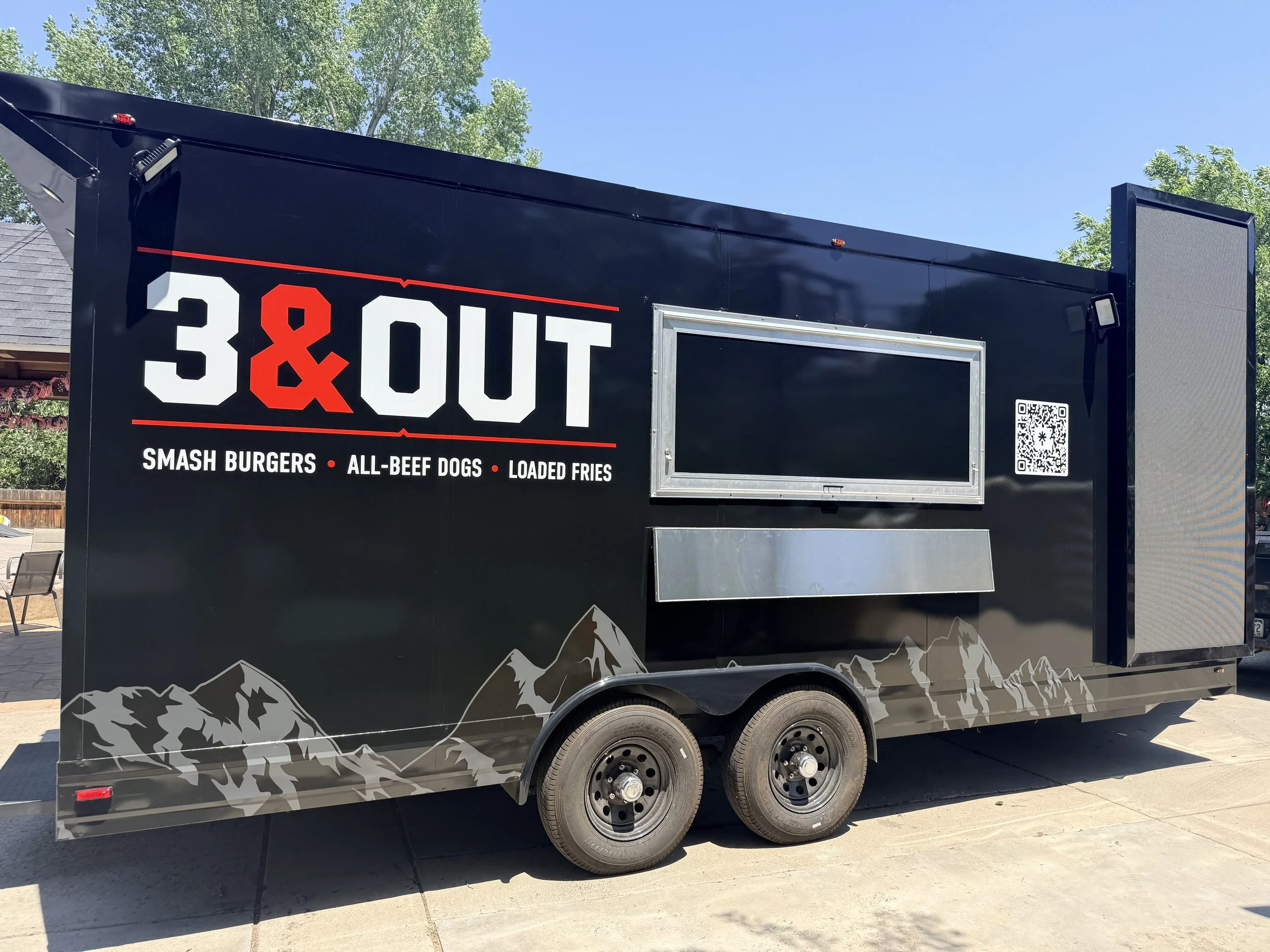 Black food truck with mountain graphics at the bottom, displaying the logo '3 & 1' in large white and red letters, and the menu items 'Smash Burgers, All-Beef Dogs, Loaded Fries' beneath the logo, parked outdoors under a clear blue sky with green tre