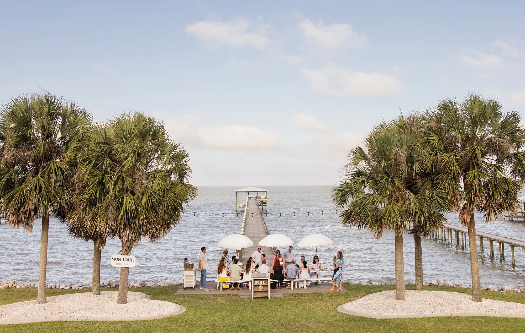 People gathered at an outdoor waterfront party under umbrellas, with a dock extending into the water and palm trees framing the scene.