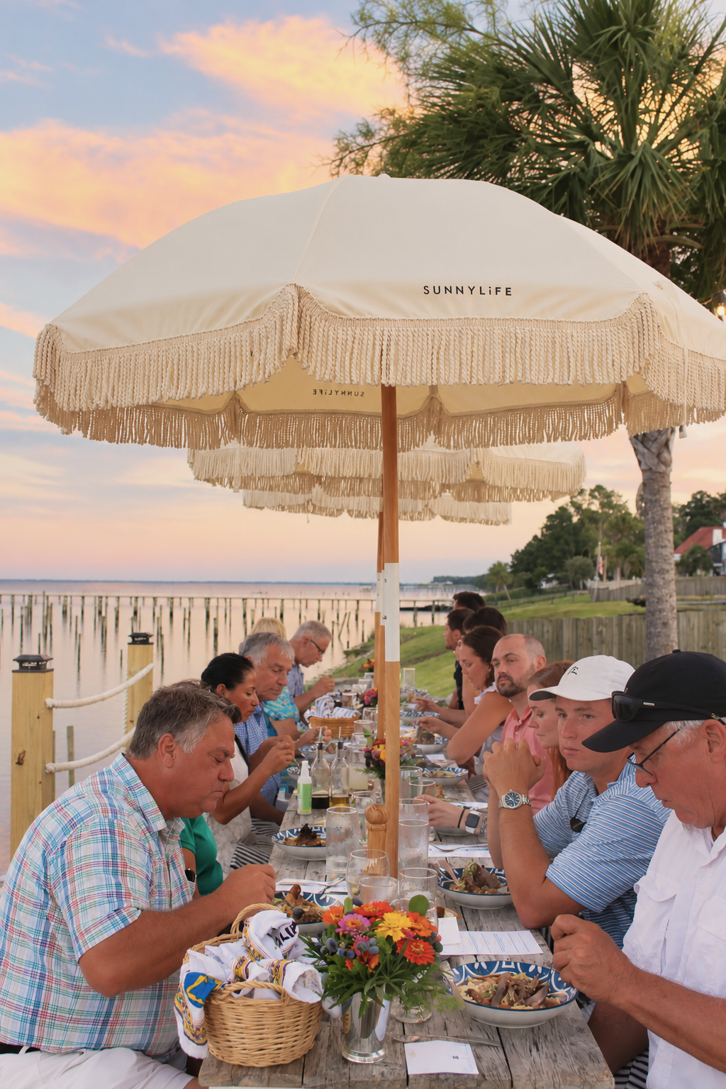 A group of people dining outdoors under large cream-colored umbrellas on a wooden pier, with a sunset sky and water in the background.