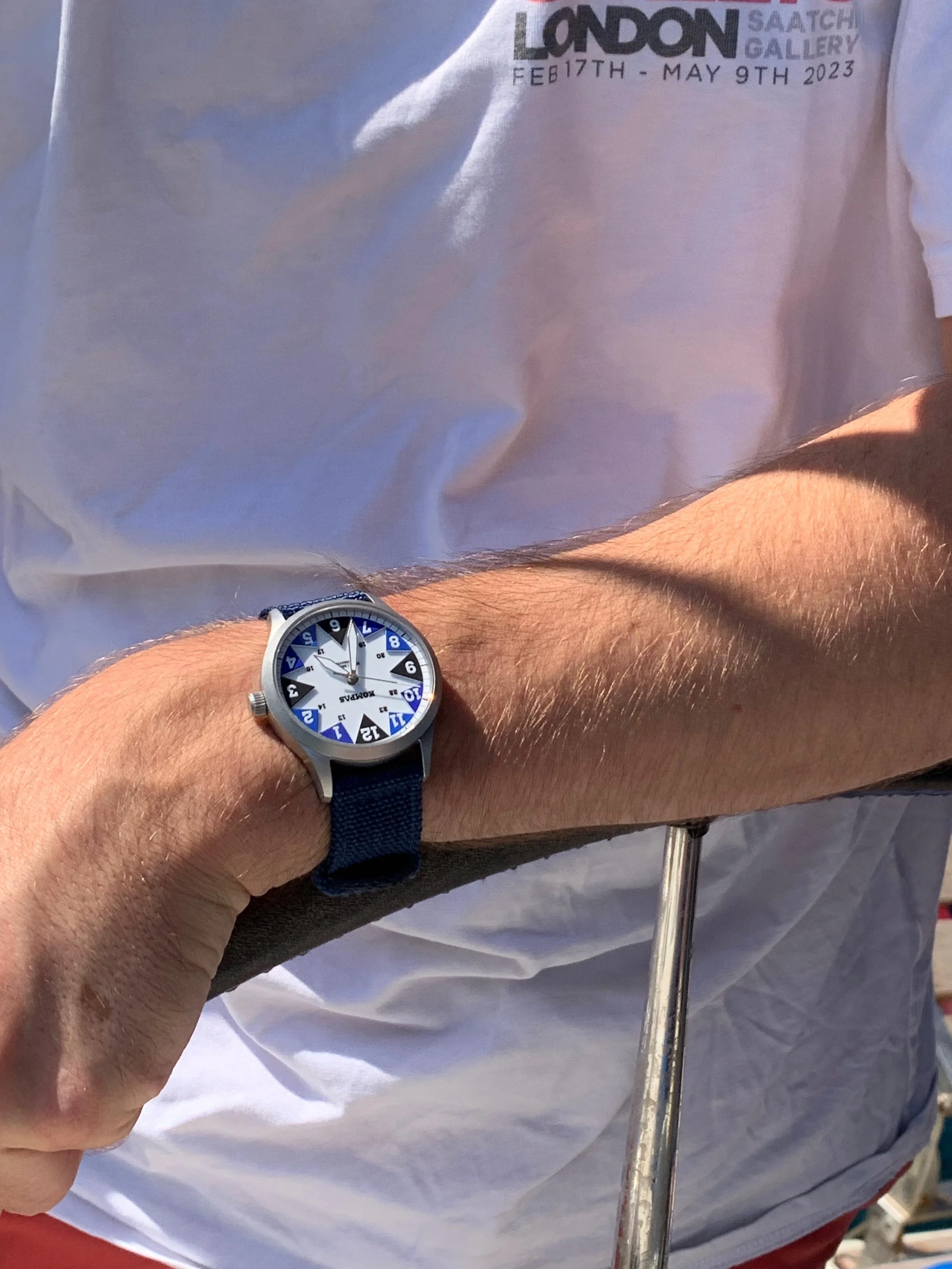 Person wearing a watch on their wrist, standing outdoors in sunlight, with a white t-shirt that has text about an exhibition in London from February 17th to May 9th, 2023.