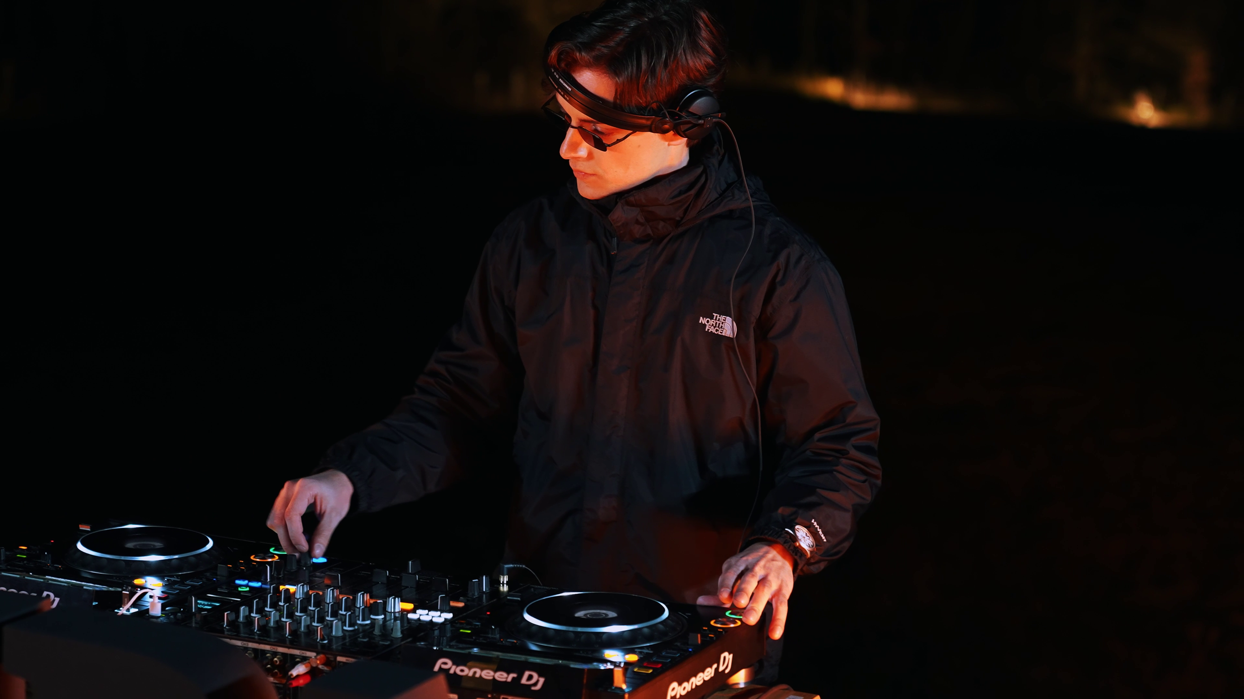 A young male DJ wearing headphones and sunglasses, dressed in a black North Face jacket, mixes music on a Pioneer DJ controller at night outdoors.