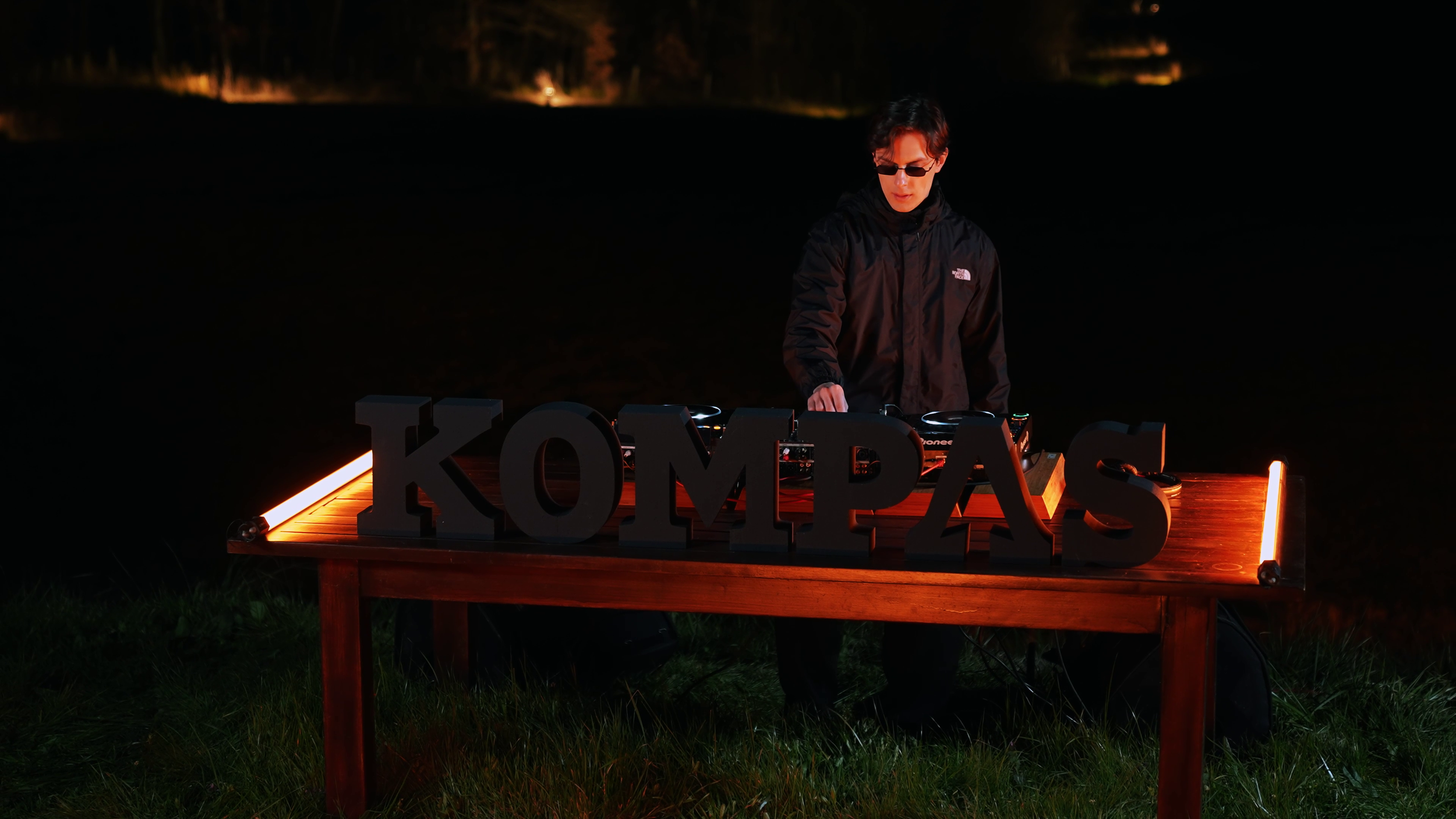 A DJ wearing black sunglasses and a black jacket performing at night behind a wooden table with illuminated Lettering spelling 'KOMPA'. The table has lights on the edges, and the background is dark with some lights in the distance.