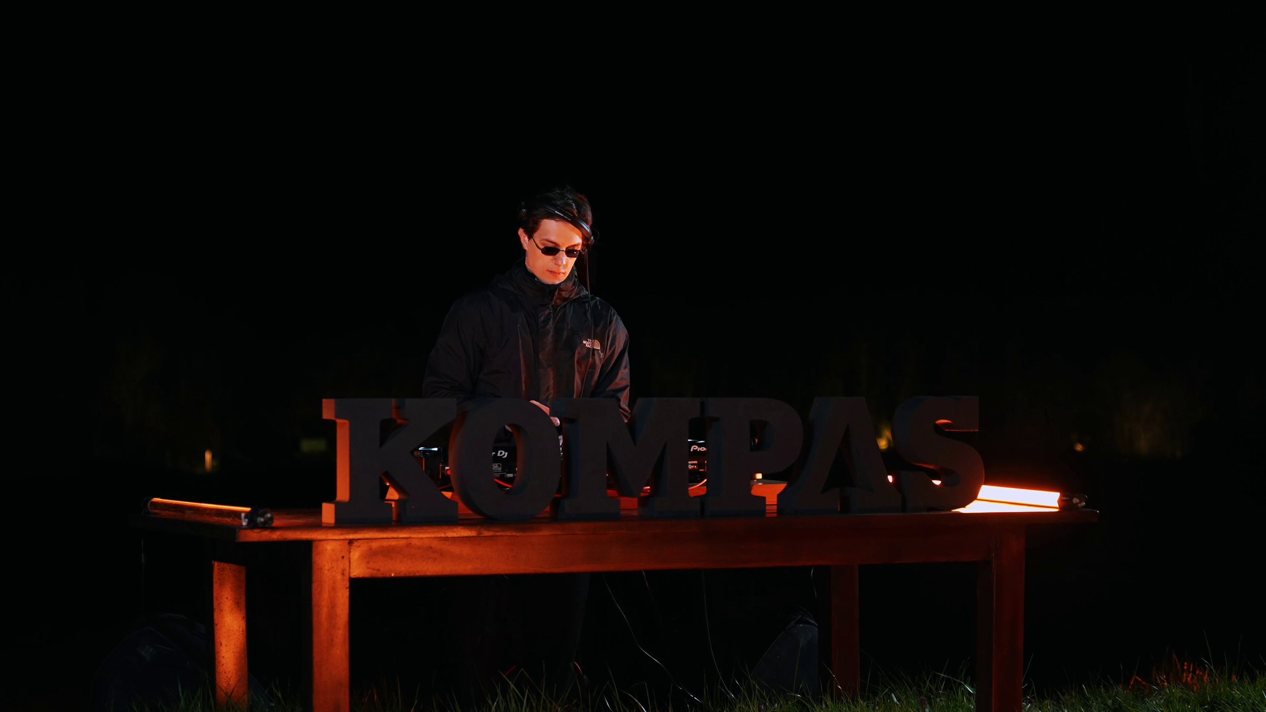 A DJ wearing sunglasses and a black jacket performing at night behind a wooden table with large black letters spelling 'KOMPAS', illuminated by orange lighting.
