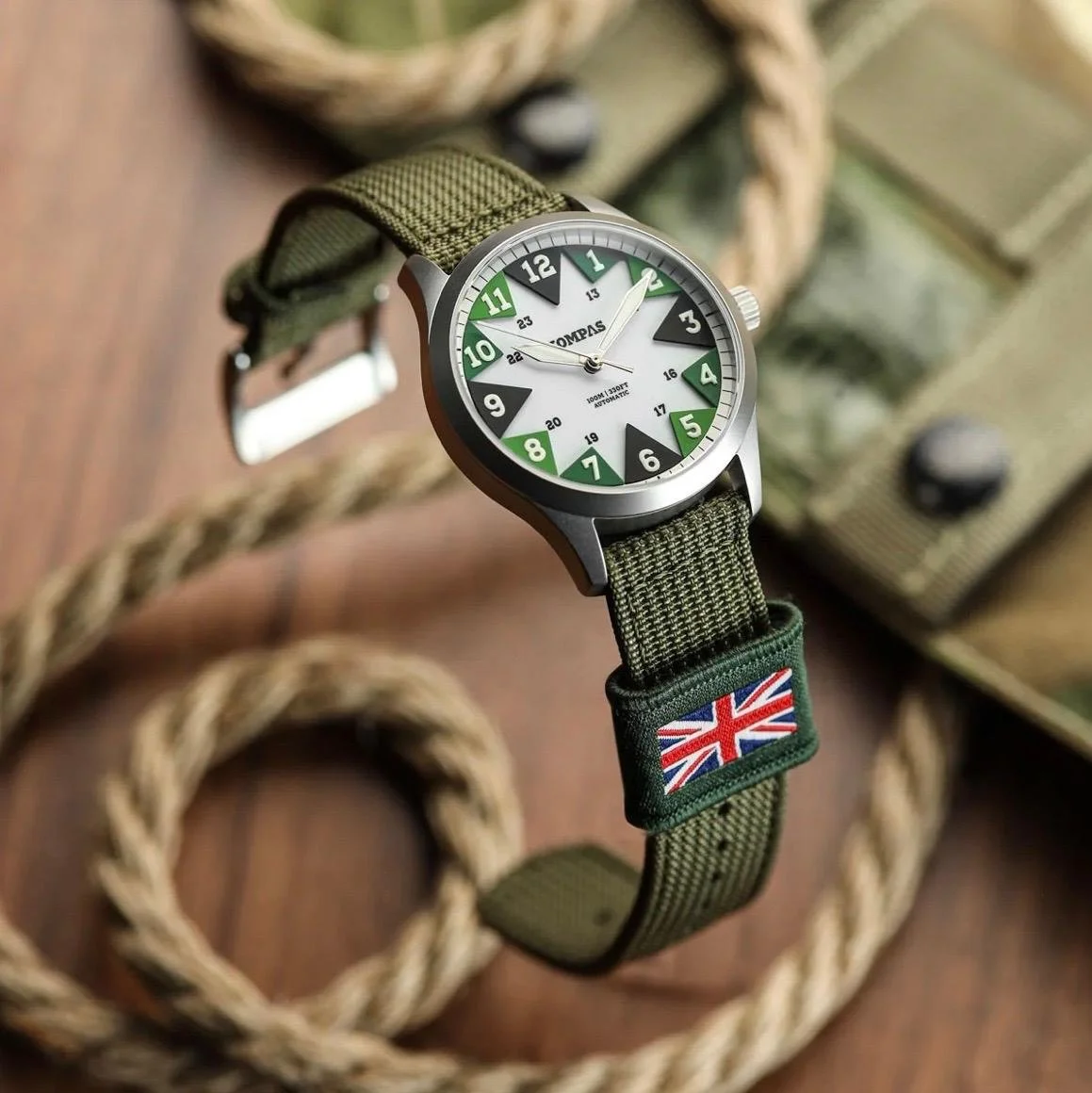A green wristwatch with a fabric strap displaying a British flag patch, placed on a wooden surface near a thick rope and a camouflage backpack.