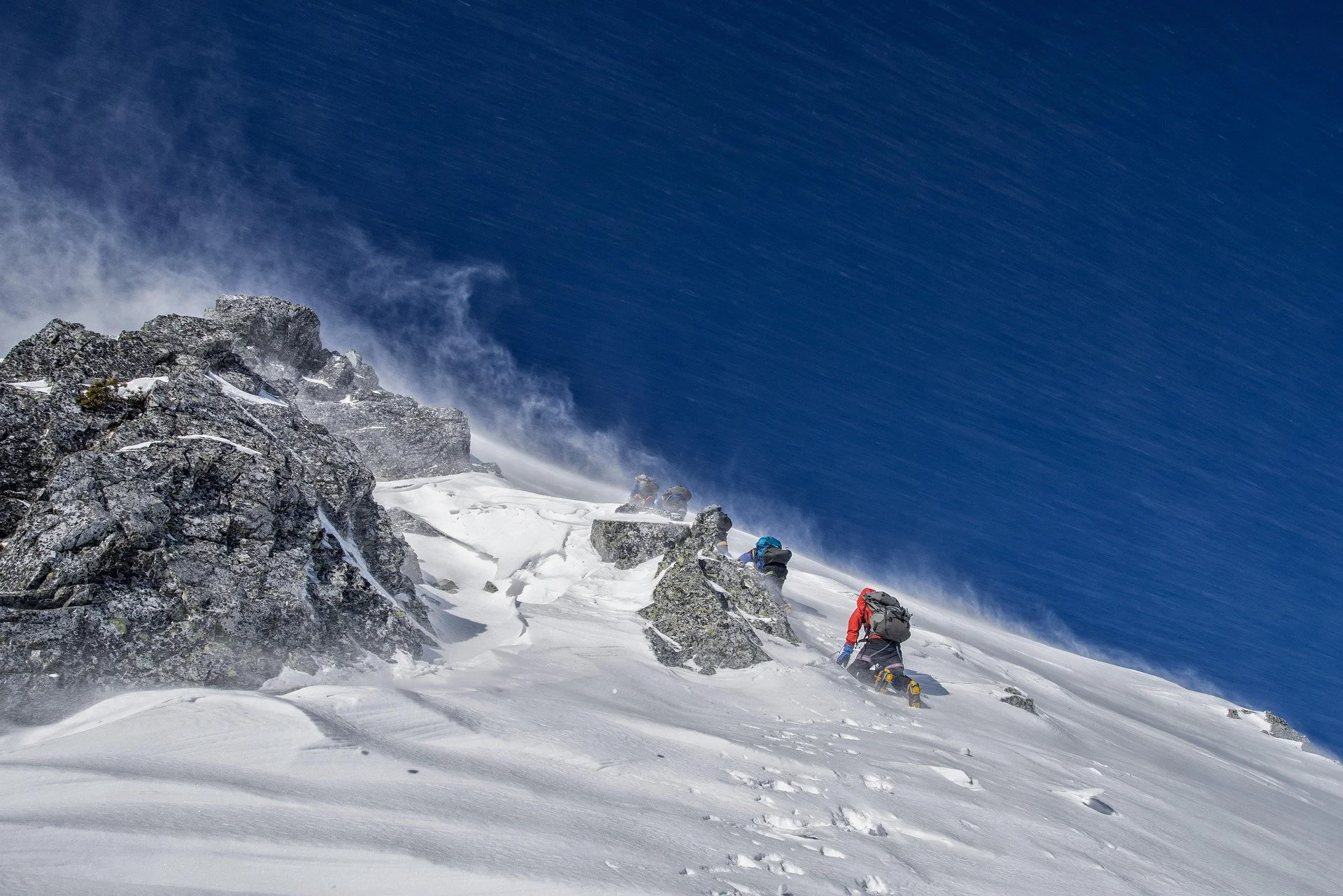 Climbers ascending a snowy, rocky mountain slope exposed to wind with snow blowing and a clear blue sky.