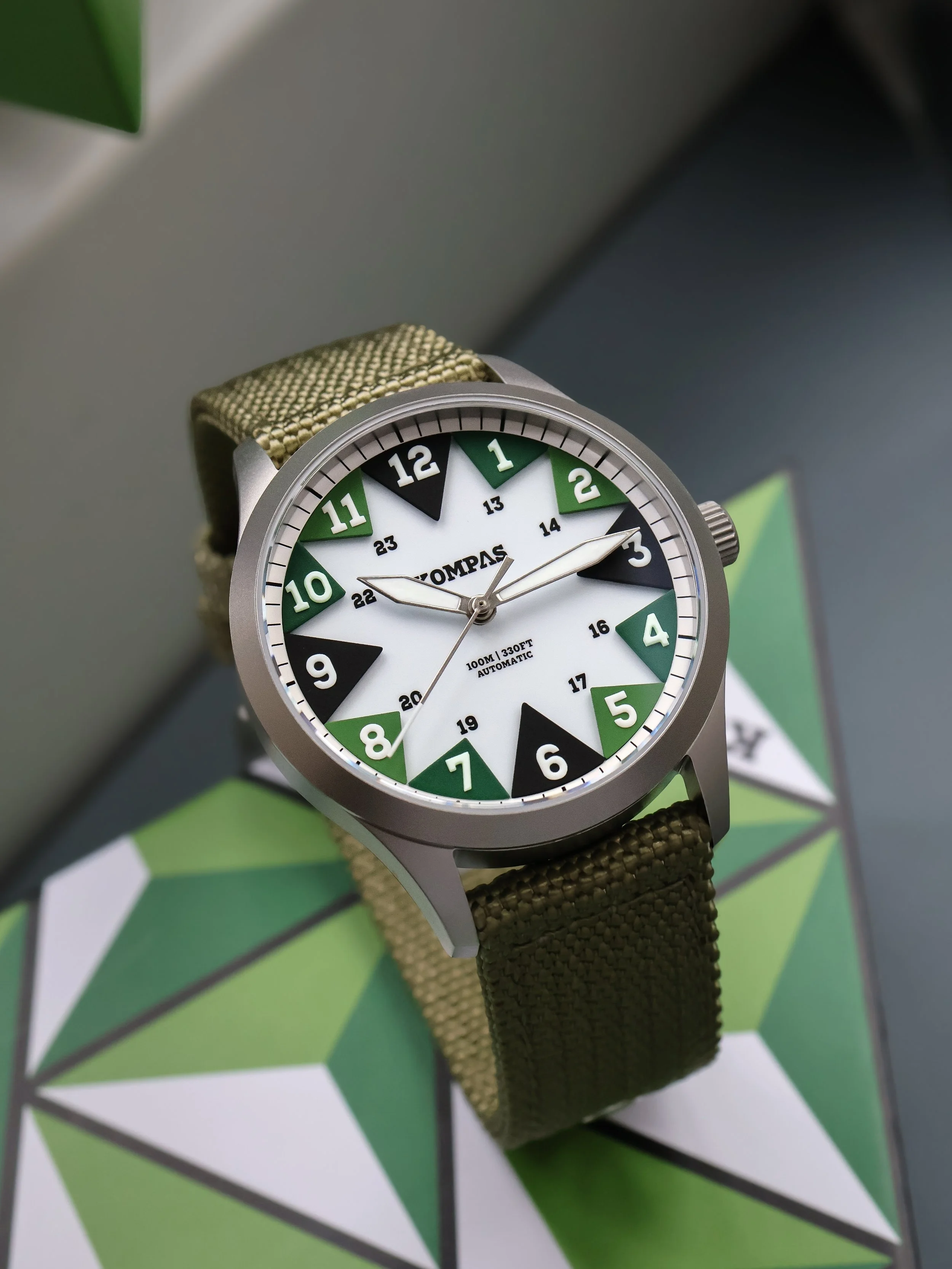 A wristwatch with a round face, green, black, and white dial, and a fabric strap, placed on a geometric-patterned surface.