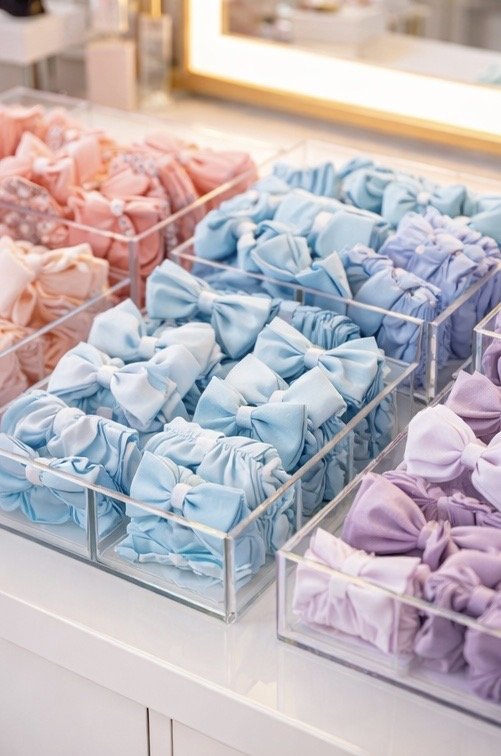 Multiple pastel-colored scrunchies and headbands arranged in clear plastic containers on a white table, with a mirror in the background.