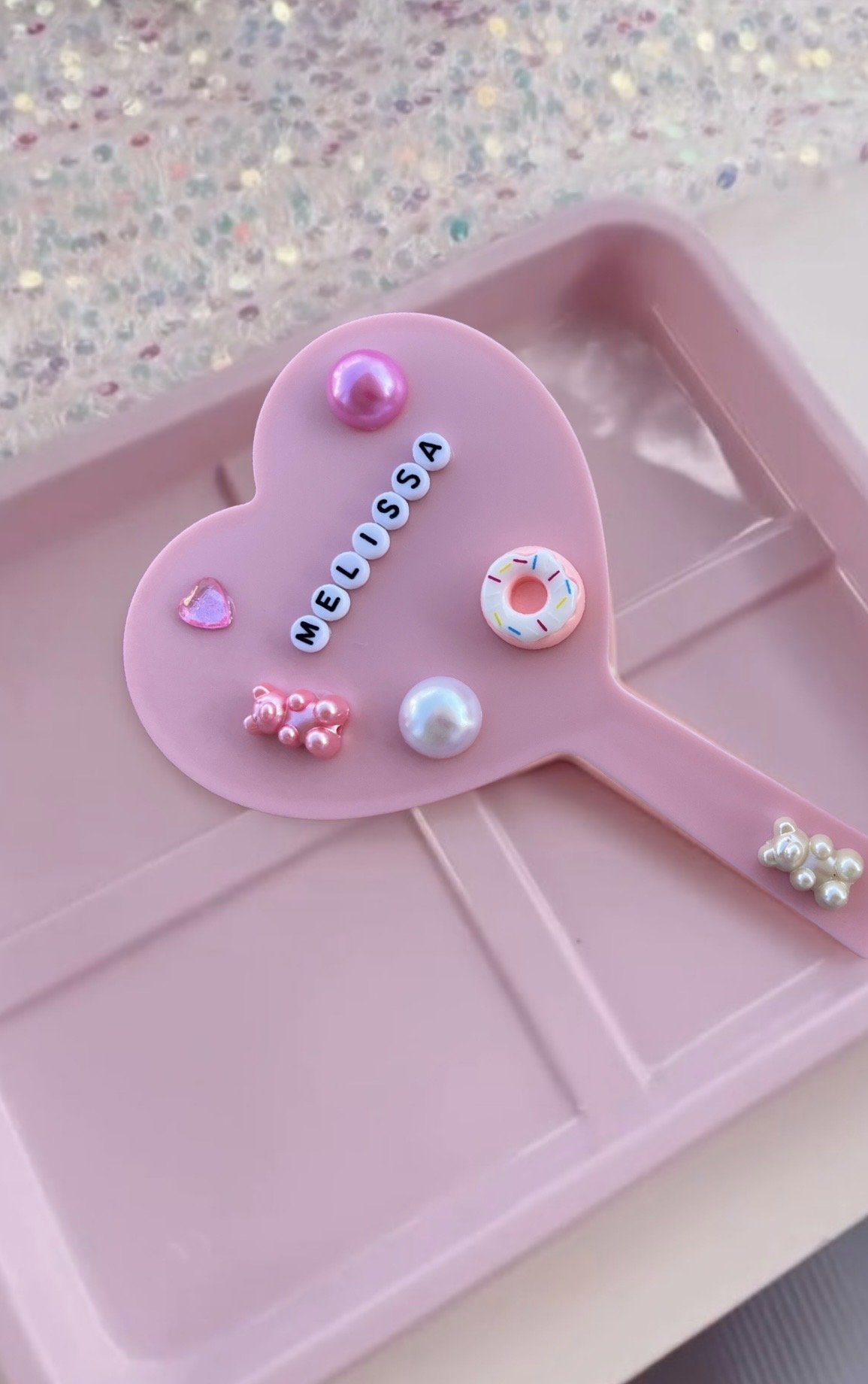 Personalized pink heart-shaped mirror decorated with pearls, beads, and charms at a Blush & Bliss by Mya craft station. Guests design their own mirrors using letter beads, donut charms, pearls, and glitter embellishments during premier kids’ spa, gla