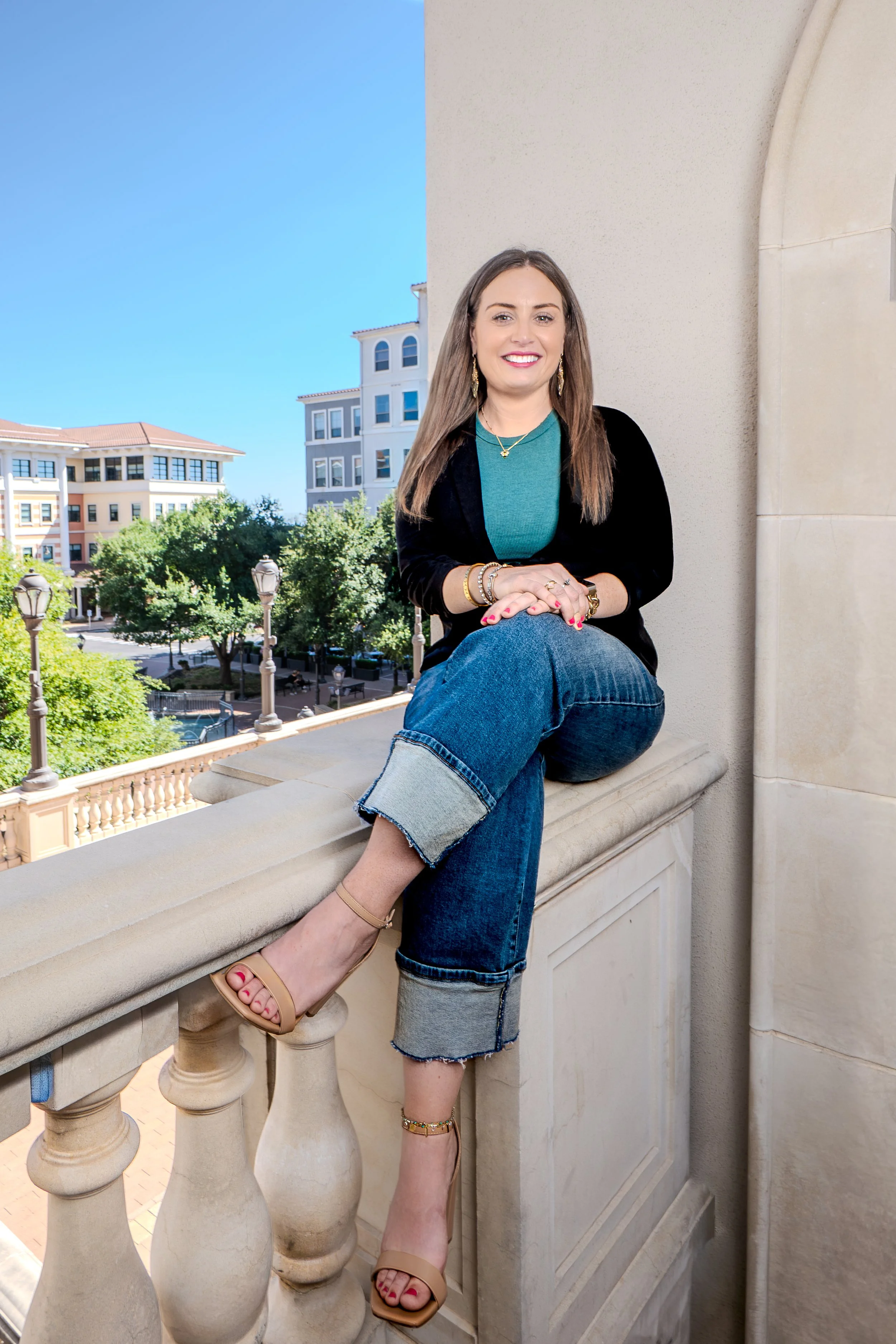 A woman sitting on a balcony ledge with city buildings and trees in the background, smiling at the camera.