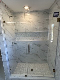 Modern glass-enclosed shower with marble tile walls and floor, metal handles, and a ceiling light fixture.