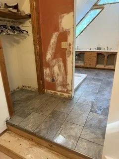 Incomplete kitchen or laundry area with exposed drywall, some flooring tiles, and a view into another room with wooden cabinets and a window.