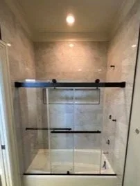Bathroom shower with glass doors, black handles, light-colored tiled walls, and a ceiling light.