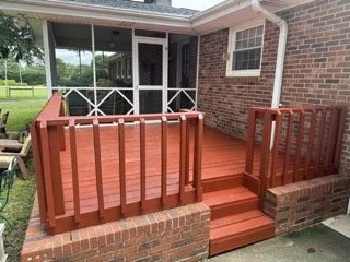 Red wooden deck with steps and railings in front of a screened porch on a brick house