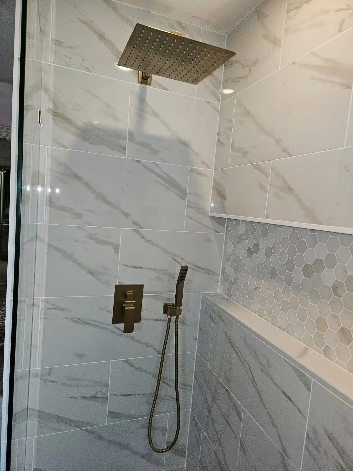A modern shower with a large rainfall showerhead, a handheld showerhead, and gold fixtures on marble and hexagonal tile walls.