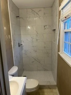 Bathroom with a walk-in shower with marble tiles, a toilet, and a window with blinds.