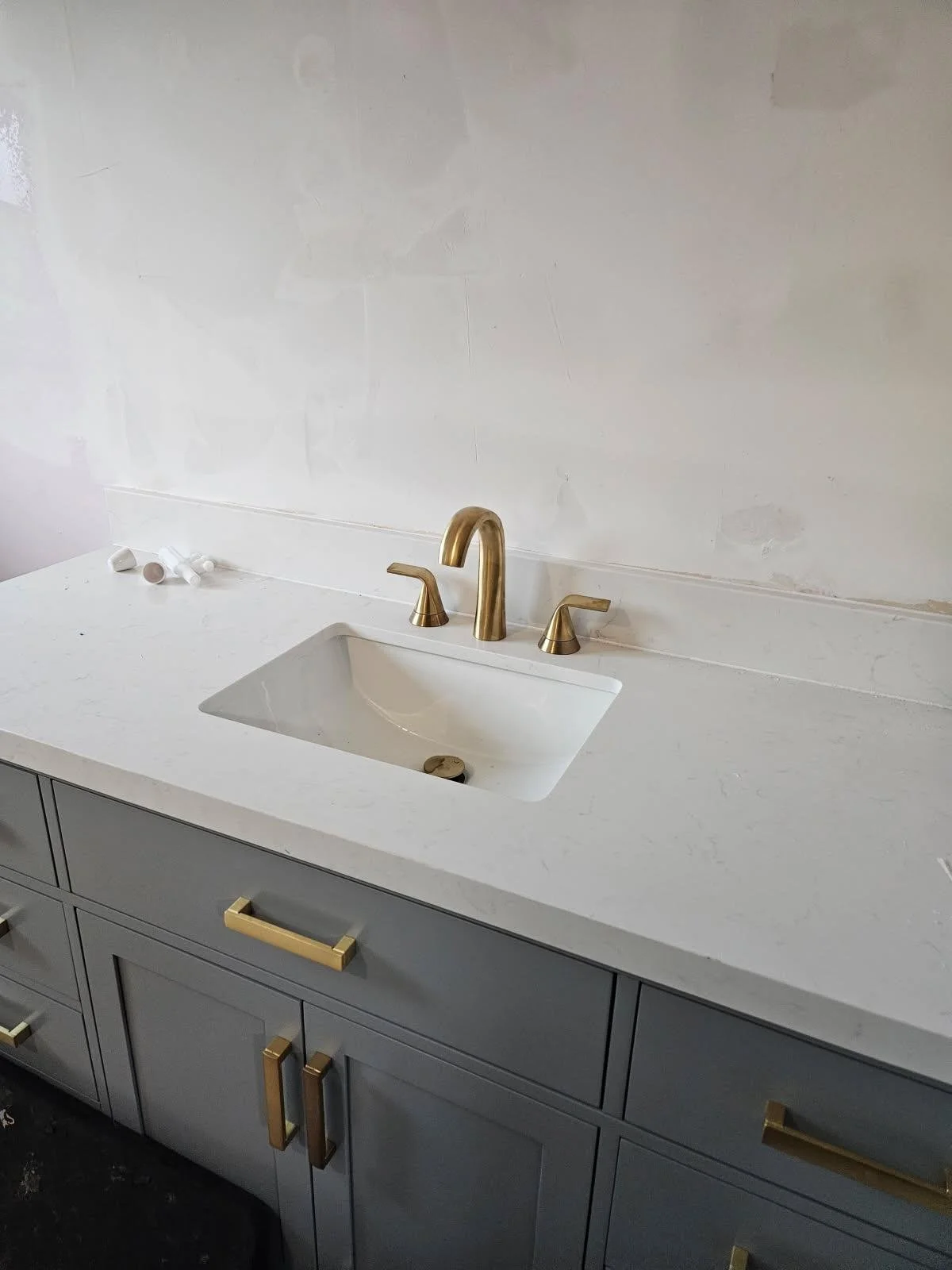 Bathroom vanity with a white countertop, a gold faucet, gray cabinets, and a soap dispenser on the left side of the counter.
