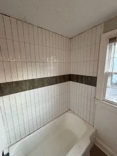 Bathroom corner with a white bathtub, tiled walls with a horizontal dark stripe, and a window with white trim.