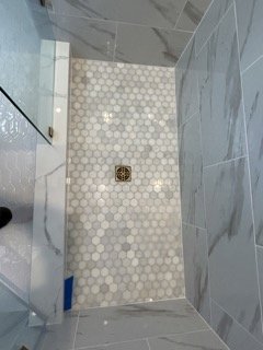 View of a bathroom shower floor with white hexagonal tiles and a drain in the center. Part of the bathroom wall with gray tiles is visible.