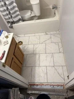 Bathroom with white tiled floor, vanity with wooden cabinet, toilet, shower with striped curtain, and a partially removed baseboard