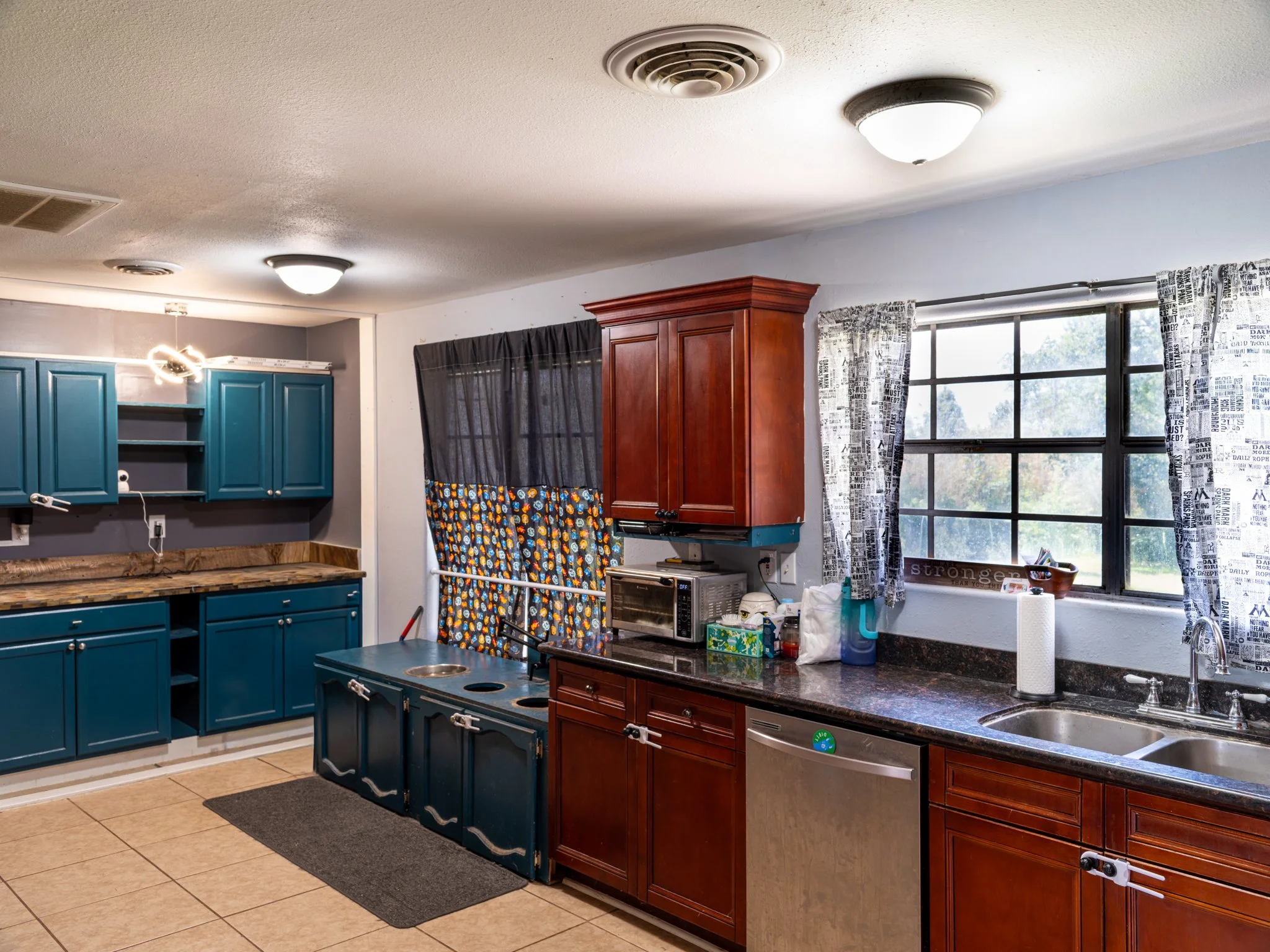 Kitchen with blue and wooden cabinets, a window with patterned curtains, and a counter with kitchen appliances.