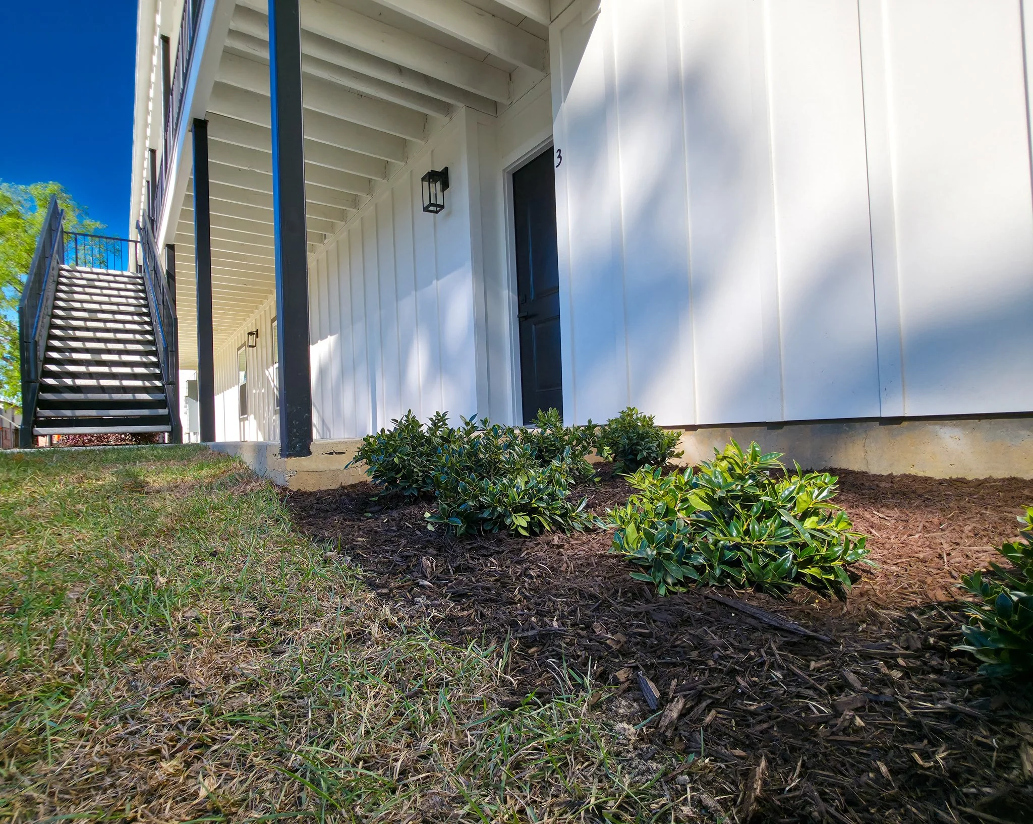 Exterior view of a modern residential building with white siding, black accents, a staircase leading to the upper level, and well-maintained landscaping with green bushes and mulch.