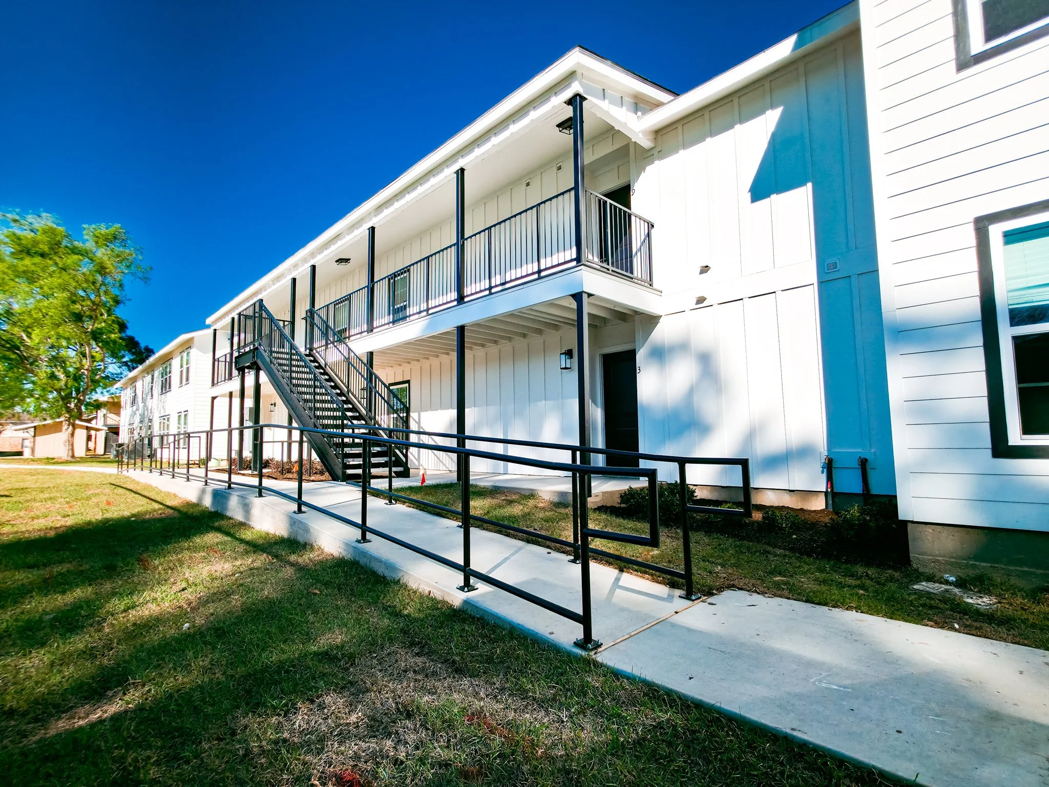 Exterior view of a modern white apartment building with a black staircase and wheelchair accessible ramp, surrounded by a grassy area and a tree.