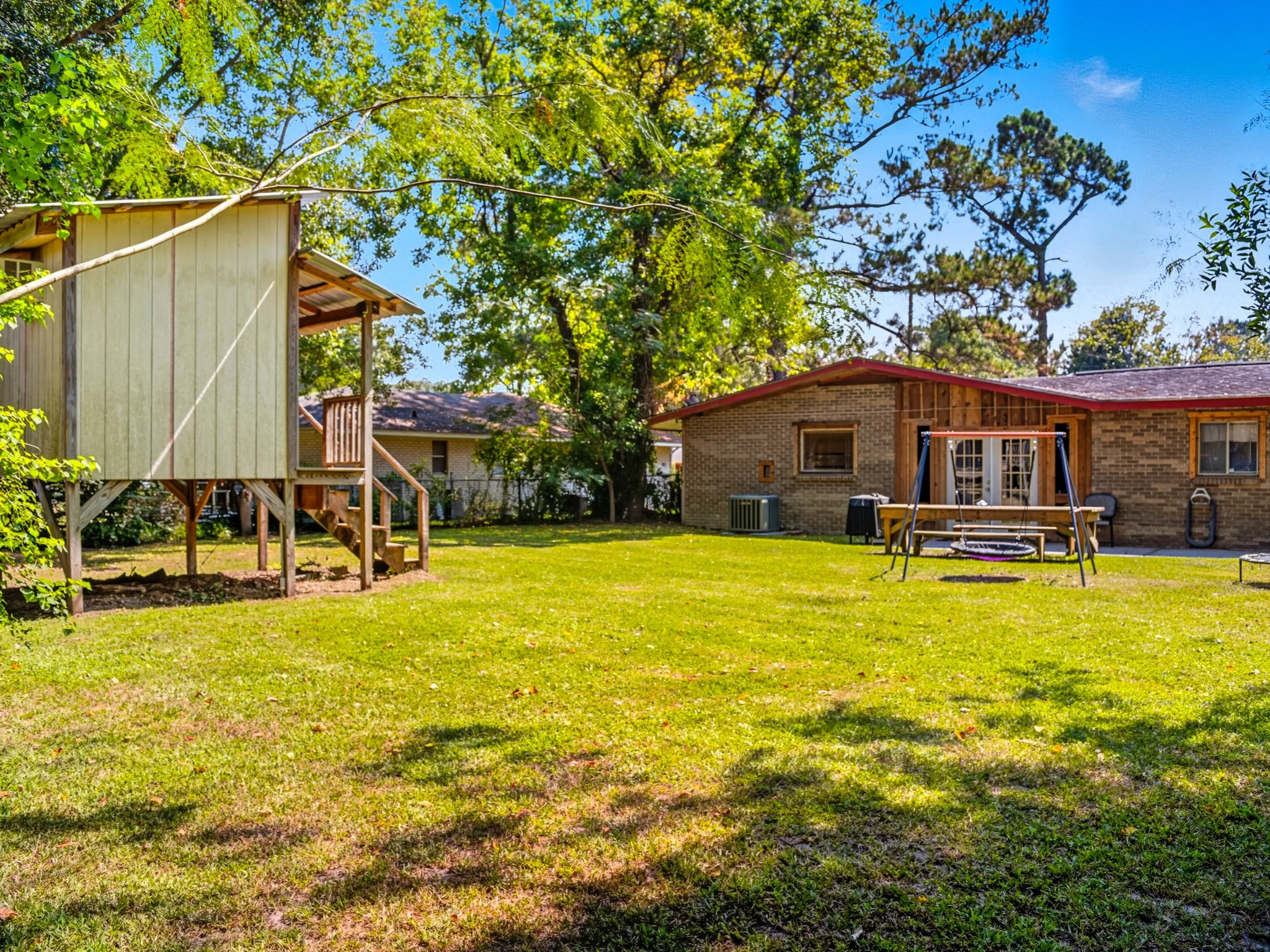 Backyard with wooden playhouse on stilts, swingset, and brick house with patio and outdoor furniture, surrounded by trees and green grass.