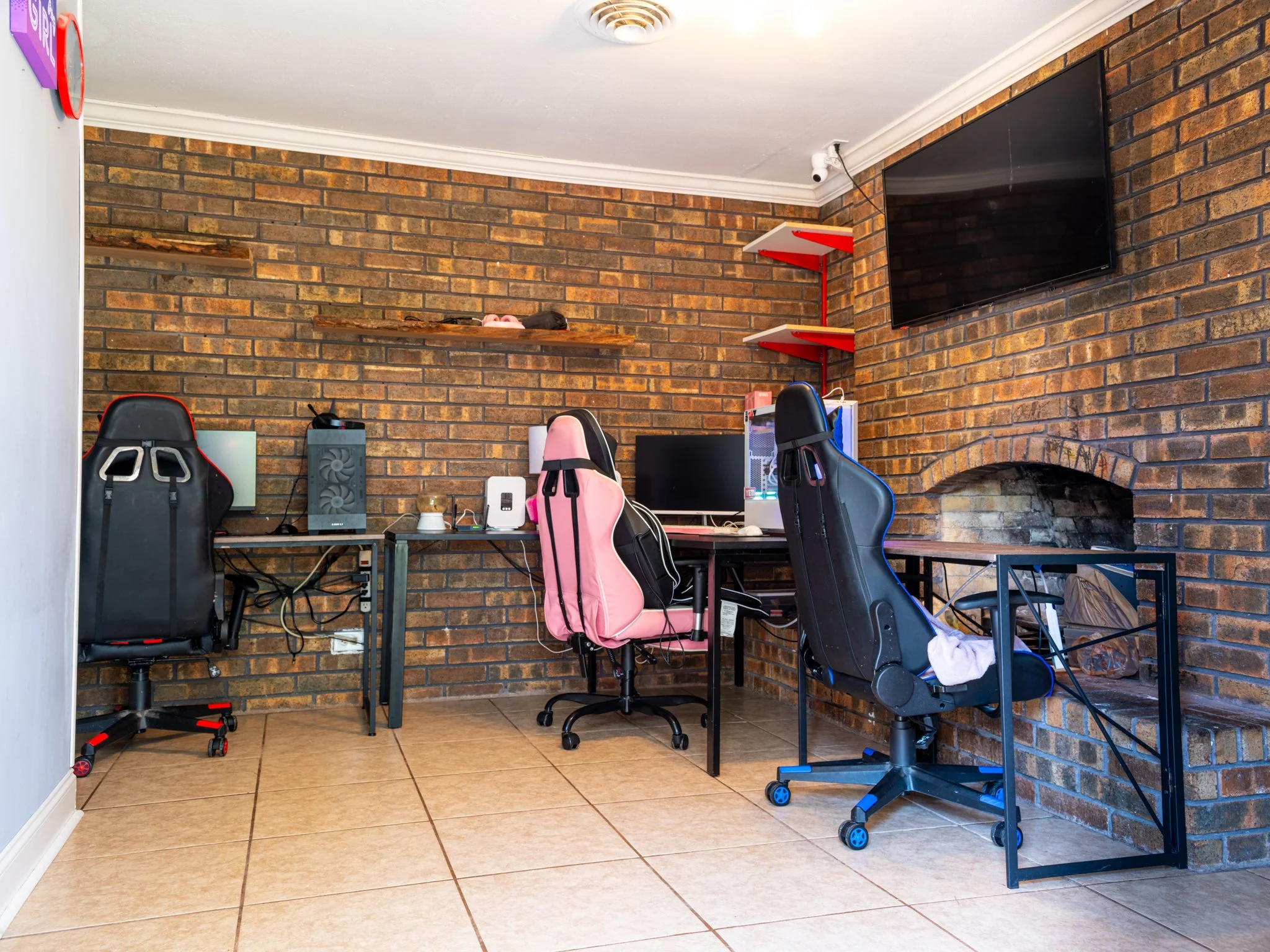 A home office with brick walls and tiled floor, featuring two desks with gaming chairs, a wall-mounted TV, and a brick fireplace.