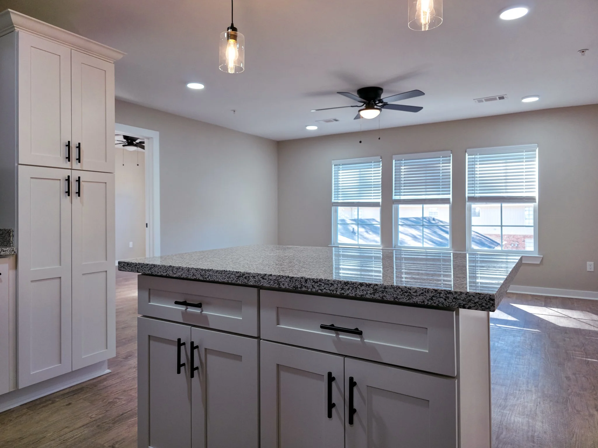 Empty modern kitchen with white cabinetry, granite island, hardwood floors, ceiling fan, and three windows with blinds allowing natural light.