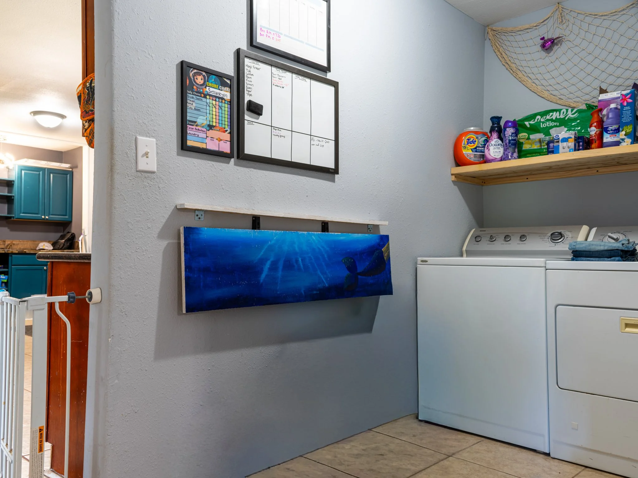 Laundry room with a washing machine, dryer, and shelves holding laundry supplies. Wall decor includes a framed chore chart, a whiteboard, and a framed list. There is a blue aquatic-themed artwork hanging on the wall.