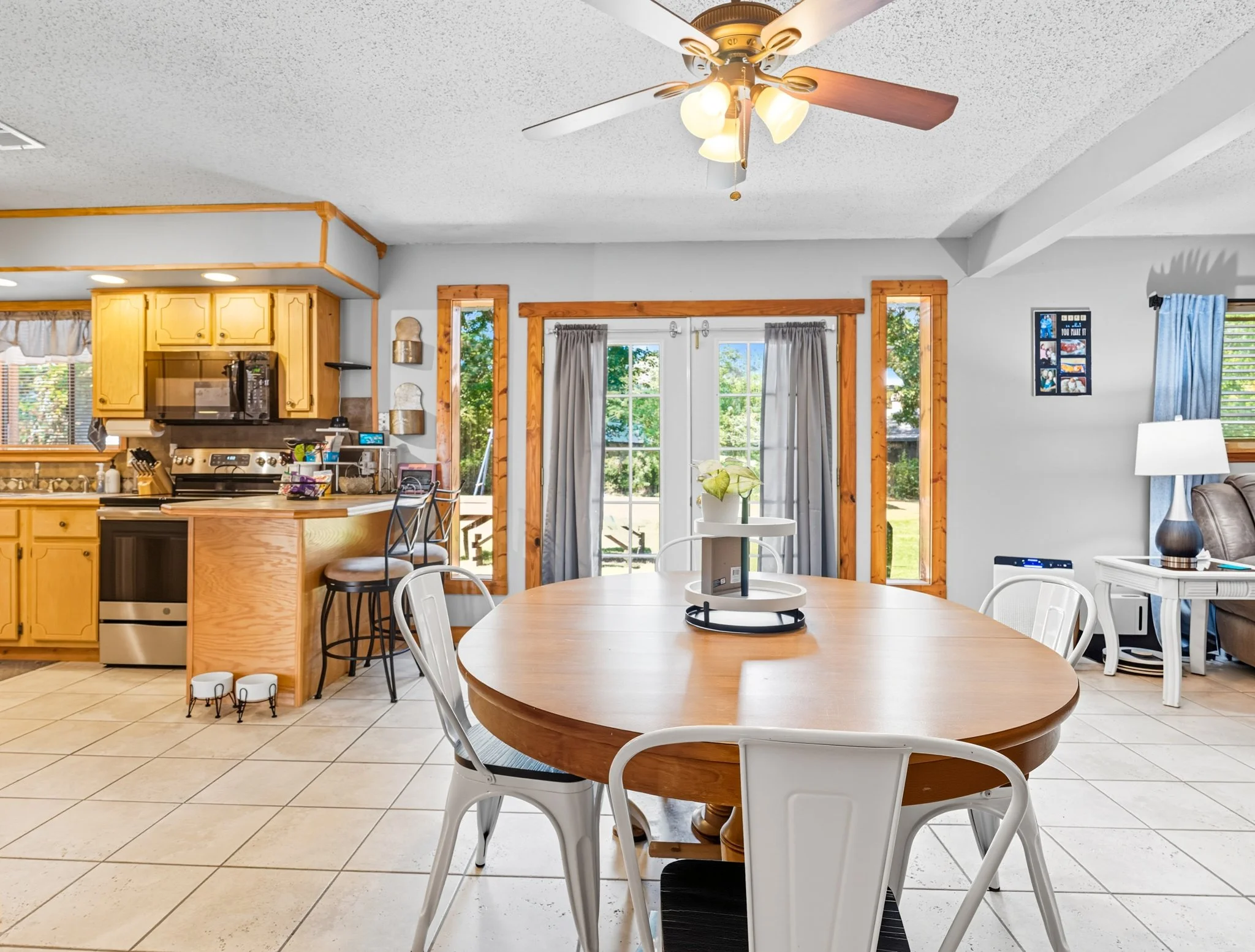 Bright kitchen and dining area with natural light streaming through glass doors and windows, a wooden dining table with white chairs, a ceiling fan with light, and kitchen cabinets with black appliances.