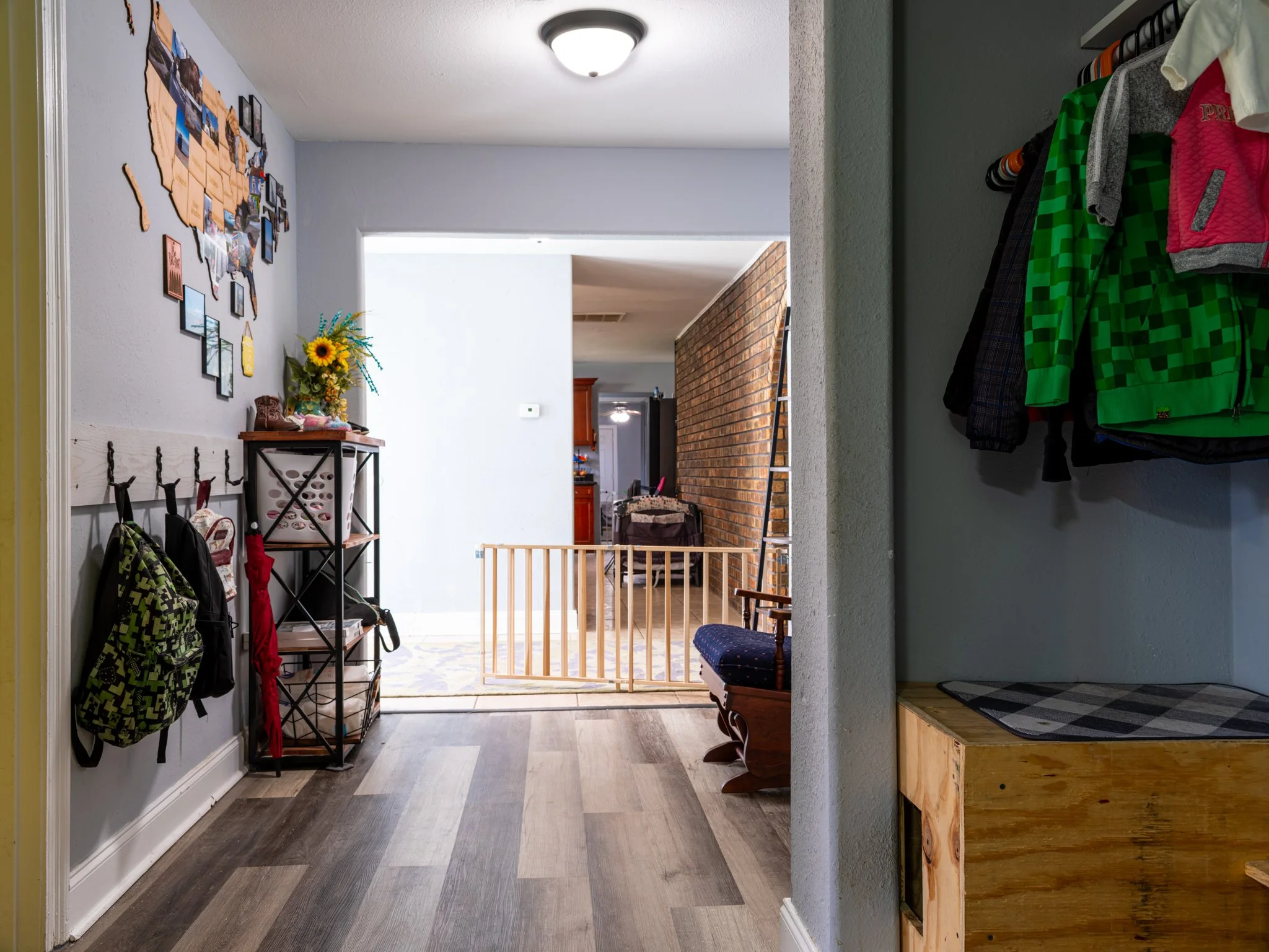 A view of a home's interior from the front door, showing an entryway with hooks on the wall holding backpacks and umbrellas, a black shelf with a sunflower arrangement, a wooden bench, and a staircase area with a safety gate, leading into the living 