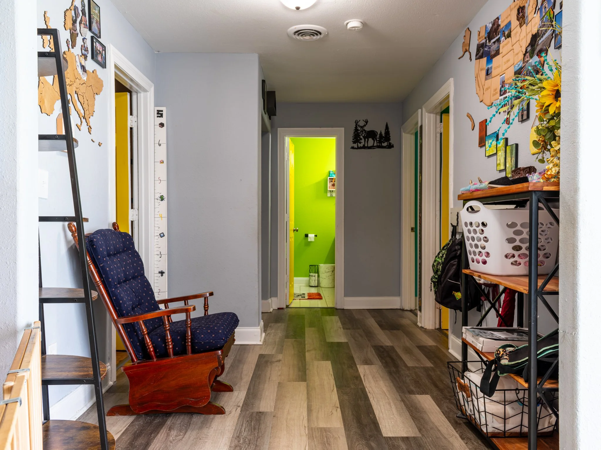 A hallway with gray walls and wood flooring, leading to a colorful bathroom with a bright green wall, decorated with various items on the walls and shelves.
