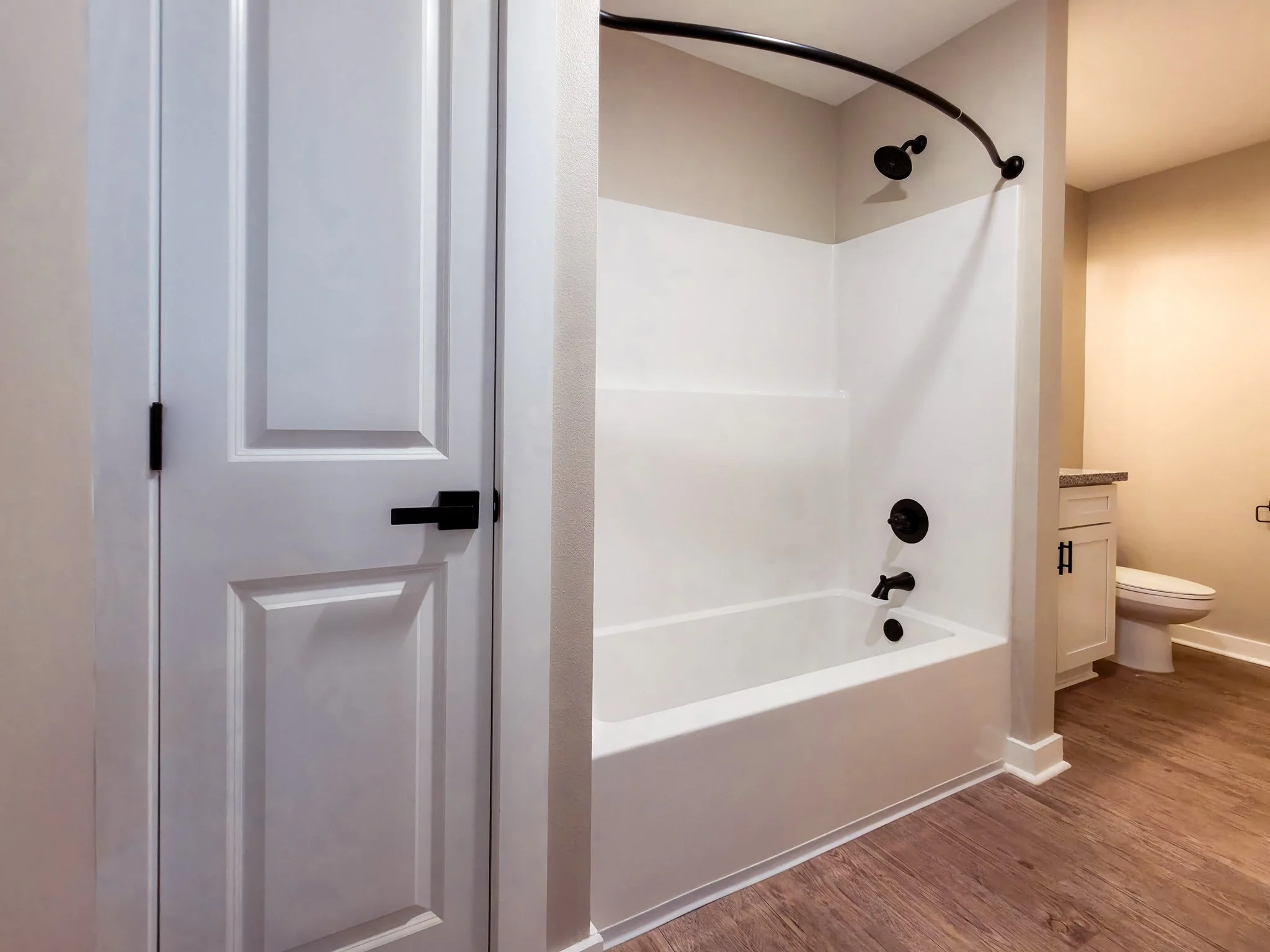Bathroom featuring a white bathtub with black fixtures, a showerhead, and a curved shower rod. To the right, a toilet and a small white cabinet with a countertop are visible. The floor is made of wood or wood-like material, and a white door with blac