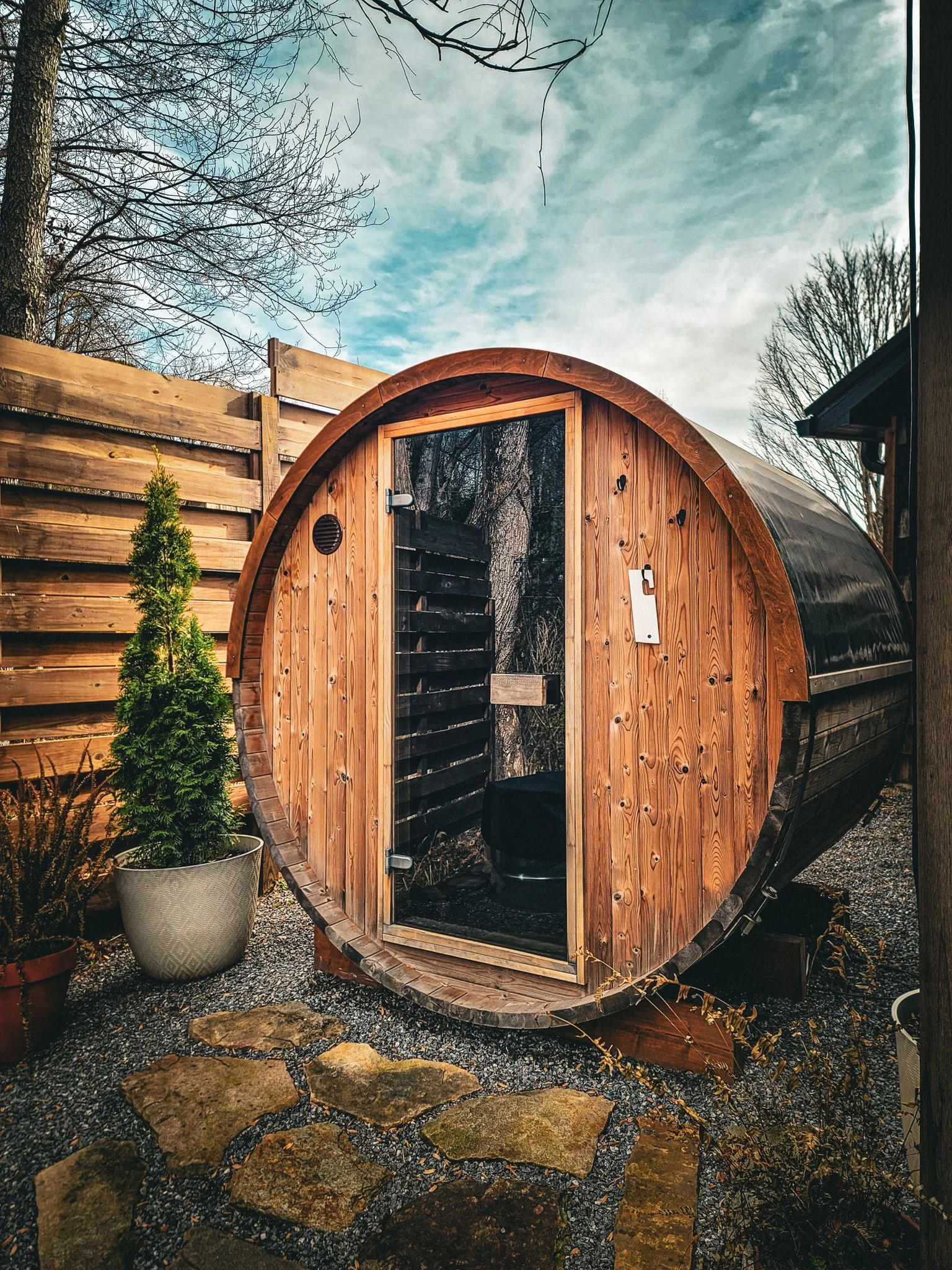 Wooden barrel-shaped sauna with glass door, situated outdoors on stone pathway, surrounded by potted plants and wooden fence, with trees and cloudy sky in the background.