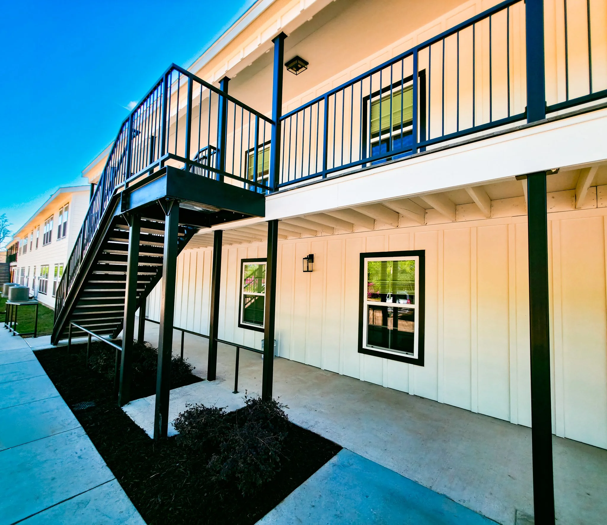 Exterior view of a two-story residential building with a metal staircase leading to the upper balcony, white siding, black window frames, and outdoor lighting, under a clear blue sky.