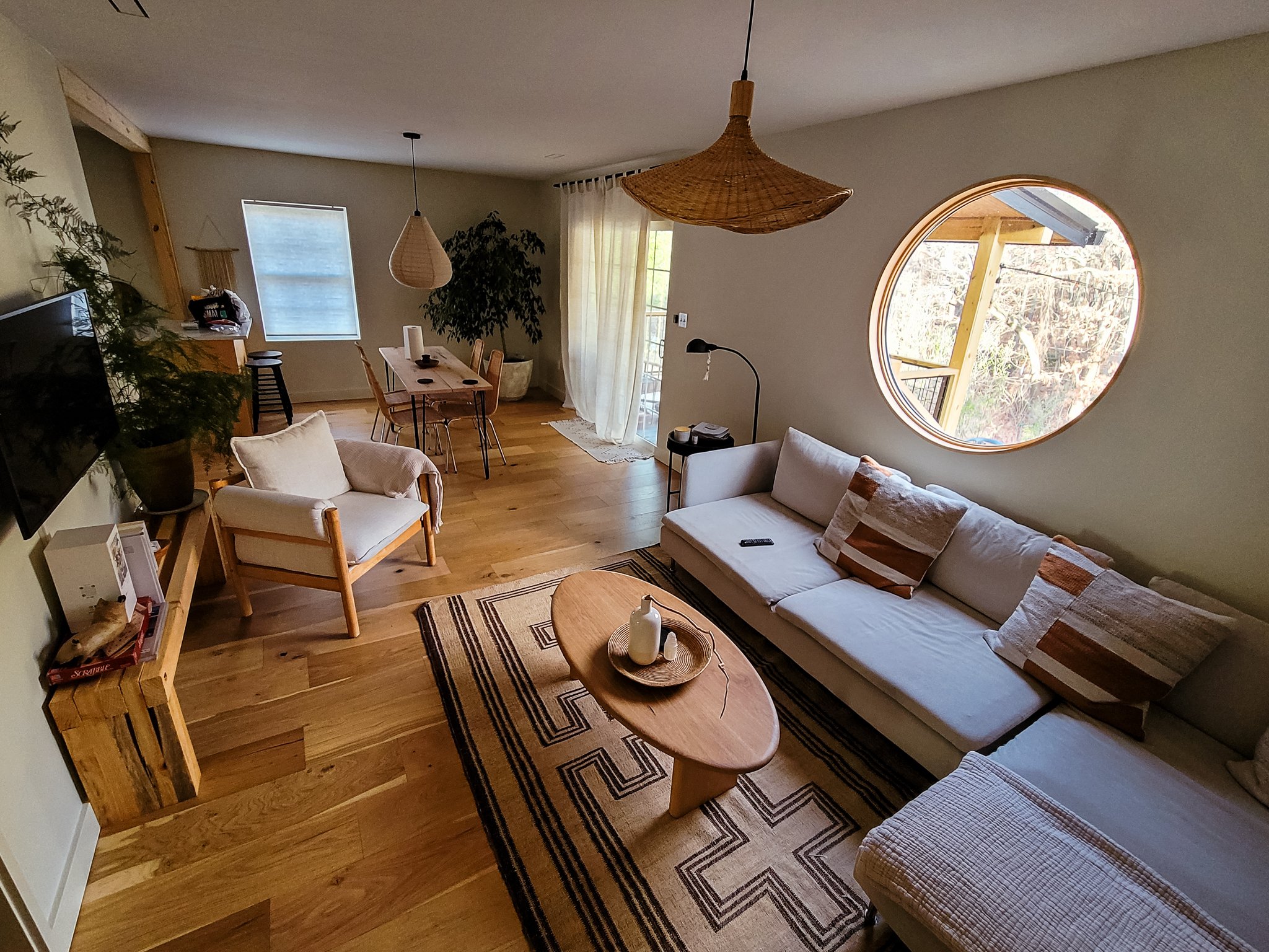 Living room with a white sectional sofa, a wooden coffee table with a bottle and a tray, a patterned area rug, a large circular window, a TV on the wall, and plants near a dining table with chairs and pendant lights, in a cozy, modern style.