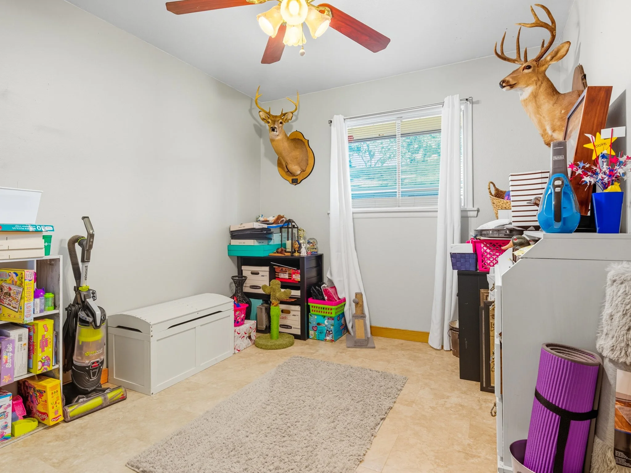 A room with beige tile floor, white walls, a window with white curtains, and two mounted deer heads on the wall. There is a ceiling fan with wooden blades and a light fixture. The room contains various storage items, toys, books, and a purple yoga ma