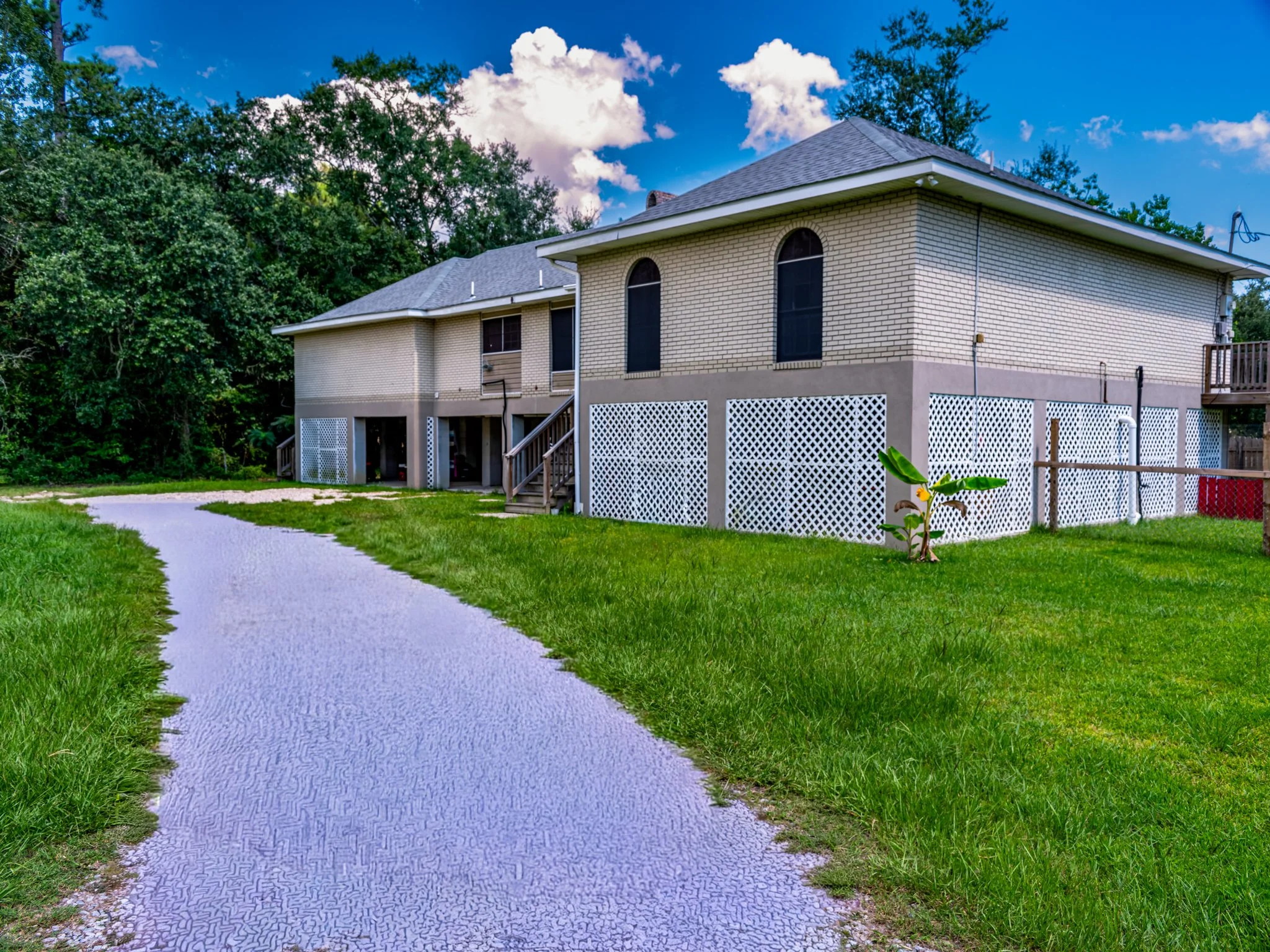 Back view of a two-story beige brick house with a gray roof, white lattice skirting, a small balcony, and a winding concrete pathway through green grass, some trees, and a partly cloudy sky.