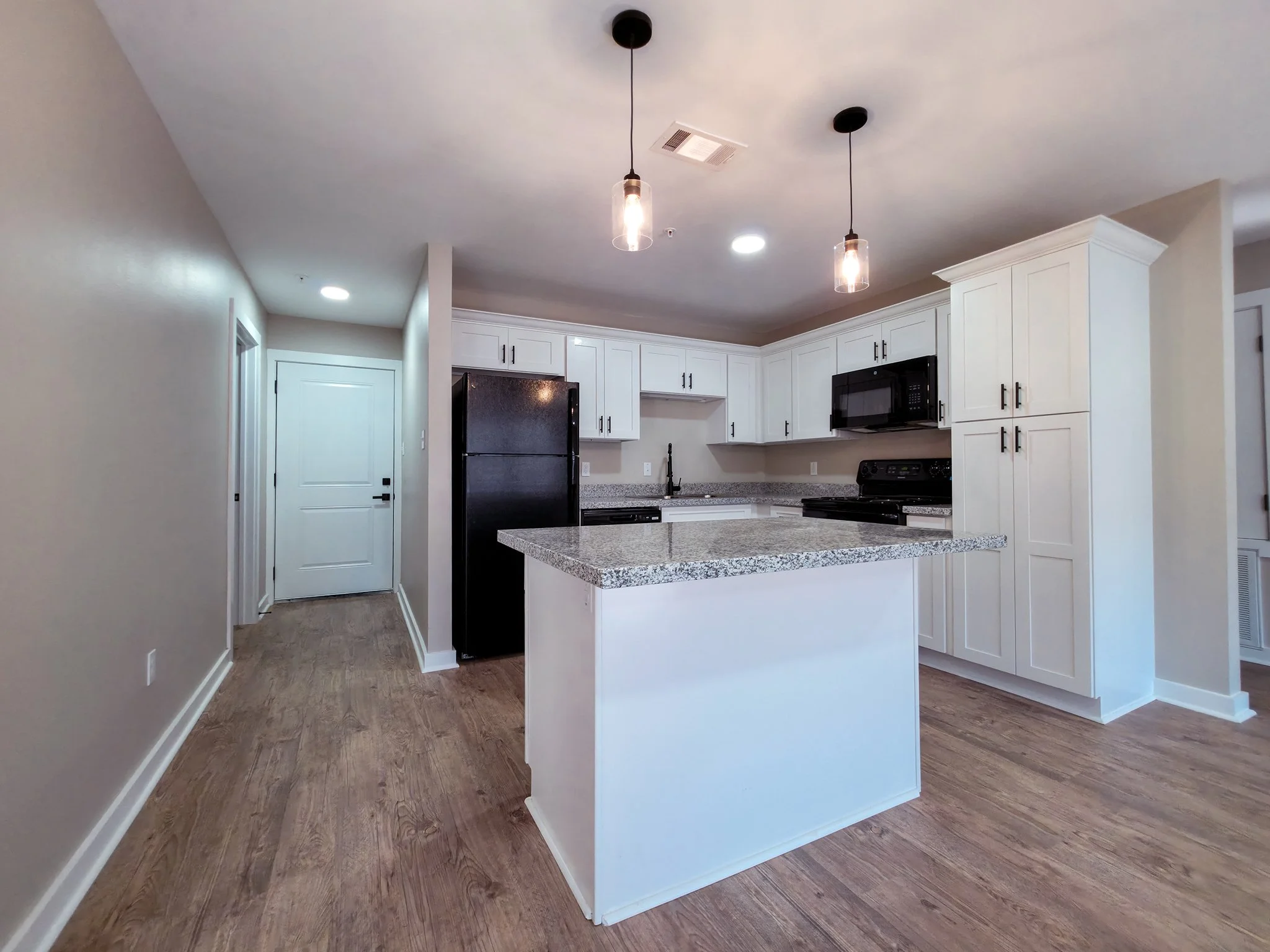 Modern kitchen with white cabinets, black appliances, granite countertops, and wooden flooring, illuminated by pendant lights.
