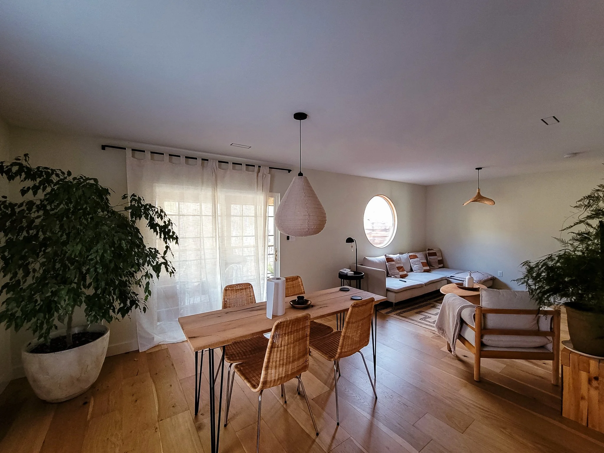 Living room with a dining table, wicker chairs, sofa, armchair, large potted plants, and natural light coming through sheer curtains and a round window.