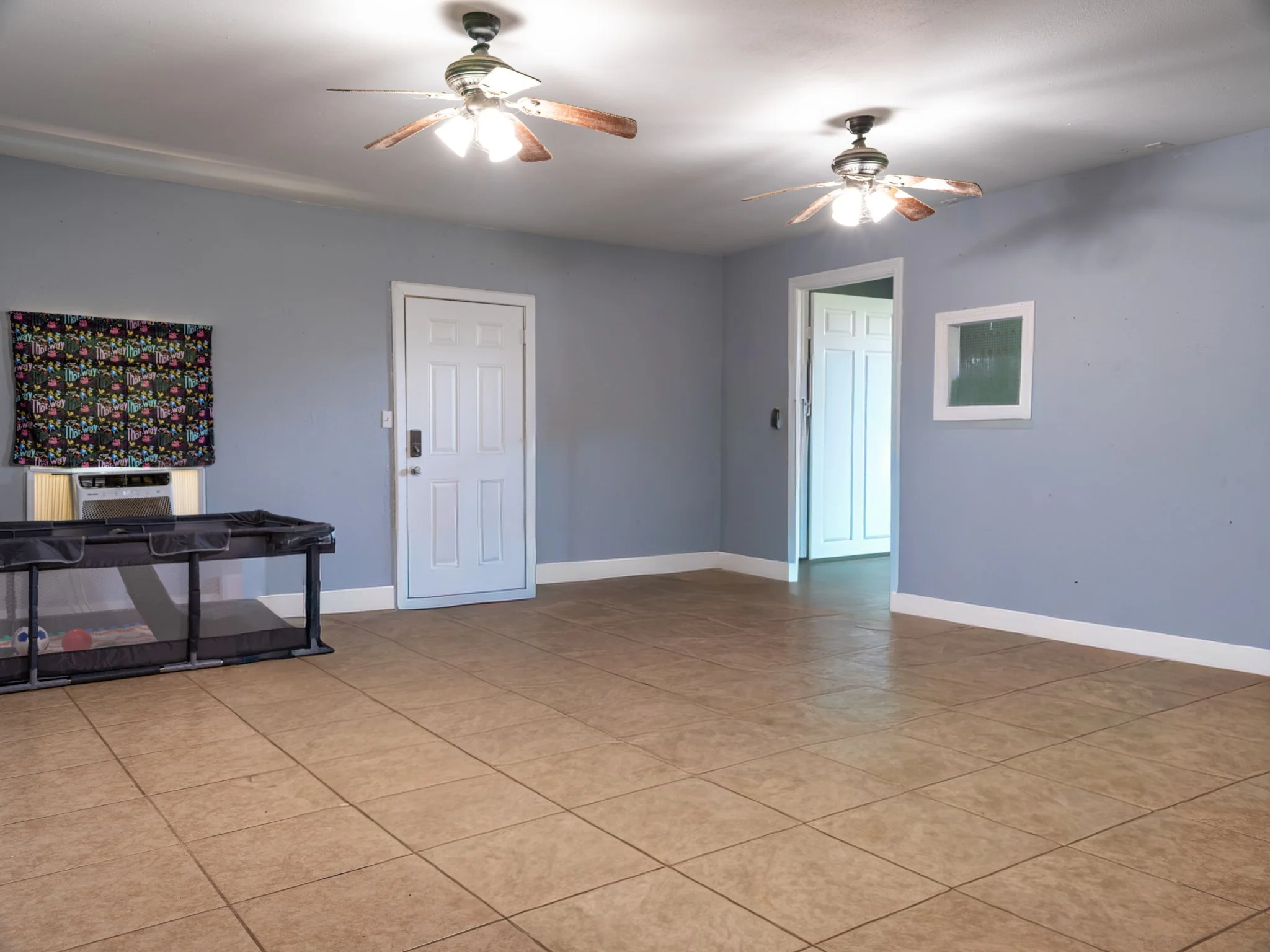 Empty living room with tiled floor, two ceiling fans with lights, a door, a window, and a small pet enclosure.