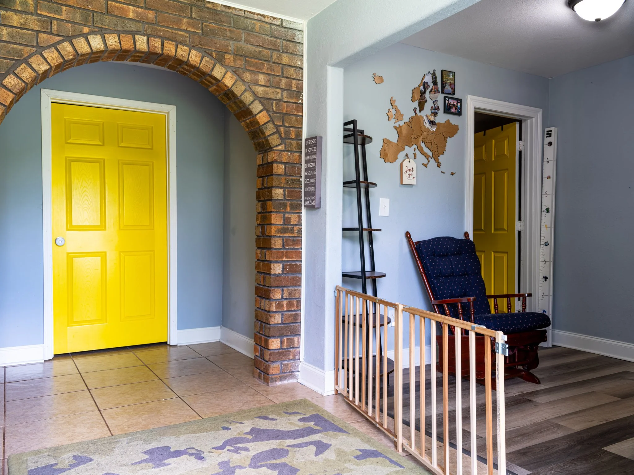 Interior view of a home entryway with a yellow door, a brick arch, a white wall with a world map and photos, a dark wooden rocking chair, and a toddler safety gate.